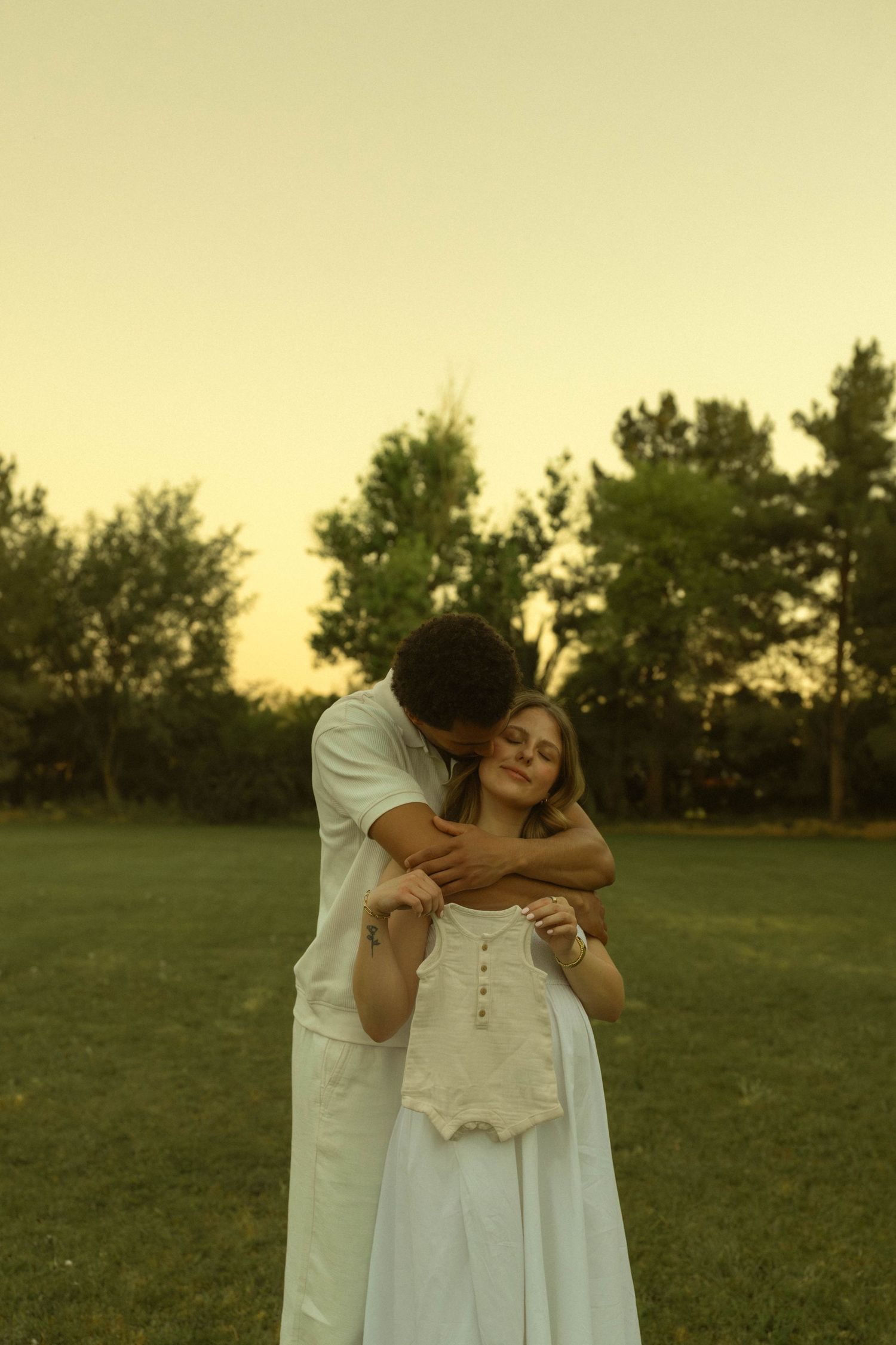 Man embracing pregnant woman holding tiny baby onesie during golden hour outdoor maternity session.
