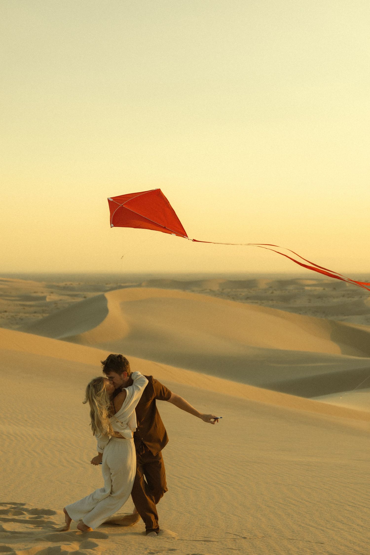 Couple kissing on sandy desert dunes at golden hour while flying a vibrant red diamond kite.