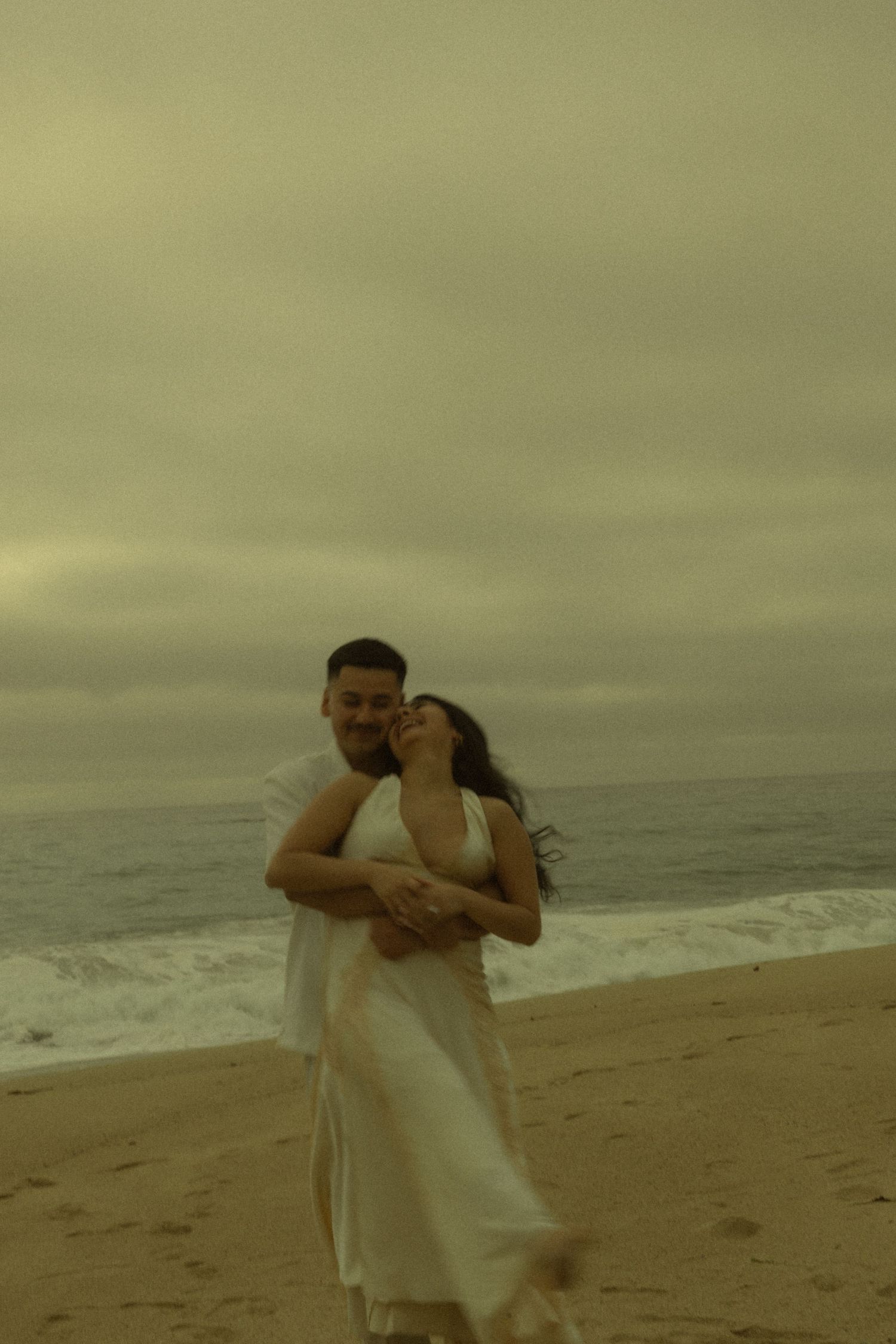 A couple embraces and laughs together on a sandy beach, wearing white outfits under a dramatic cloudy sky.
