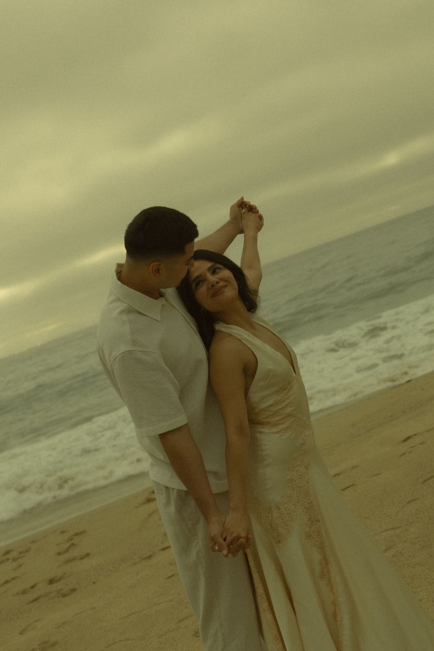 A couple dances romantically on a sandy beach at dusk, the woman in a flowing gown with waves and cloudy skies behind them.