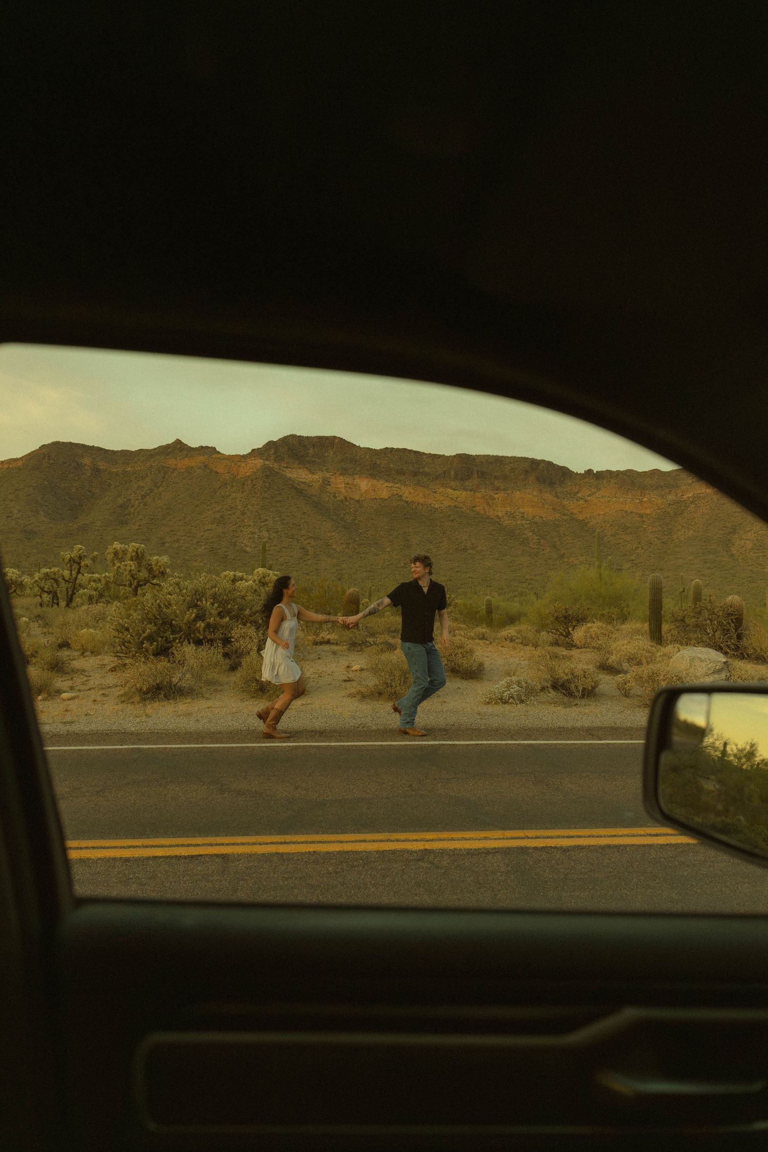 Couple holding hands crossing a desert road viewed through a car window with mountains in background.