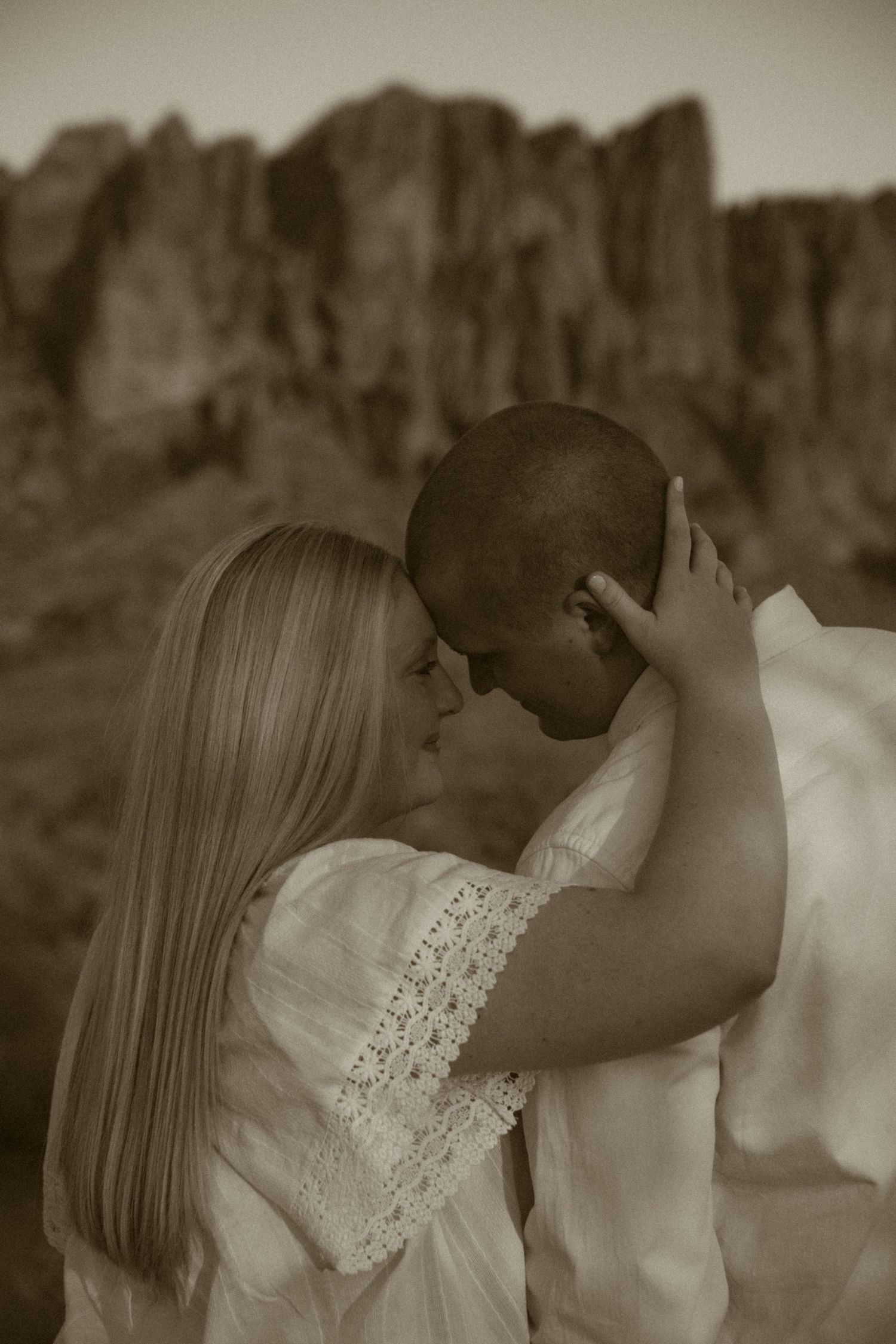 A romantic couple shares an intimate moment outdoors, with rocky cliffs in the background, in a sepia-toned portrait.
