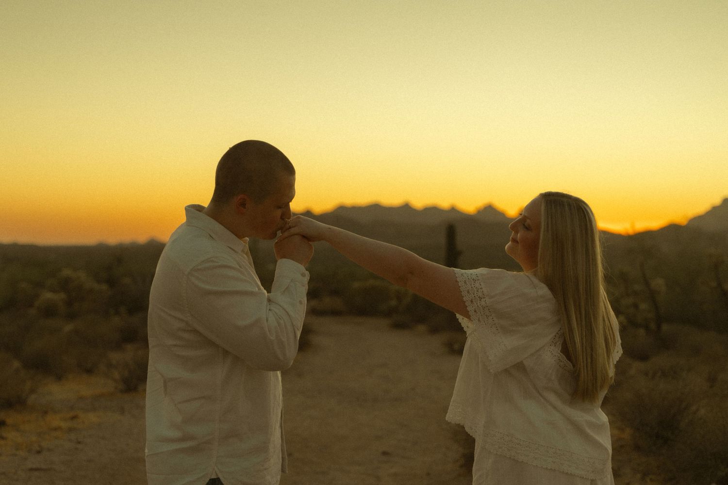 Man kissing woman's hand during a romantic desert sunset engagement photo session with golden sky backdrop.