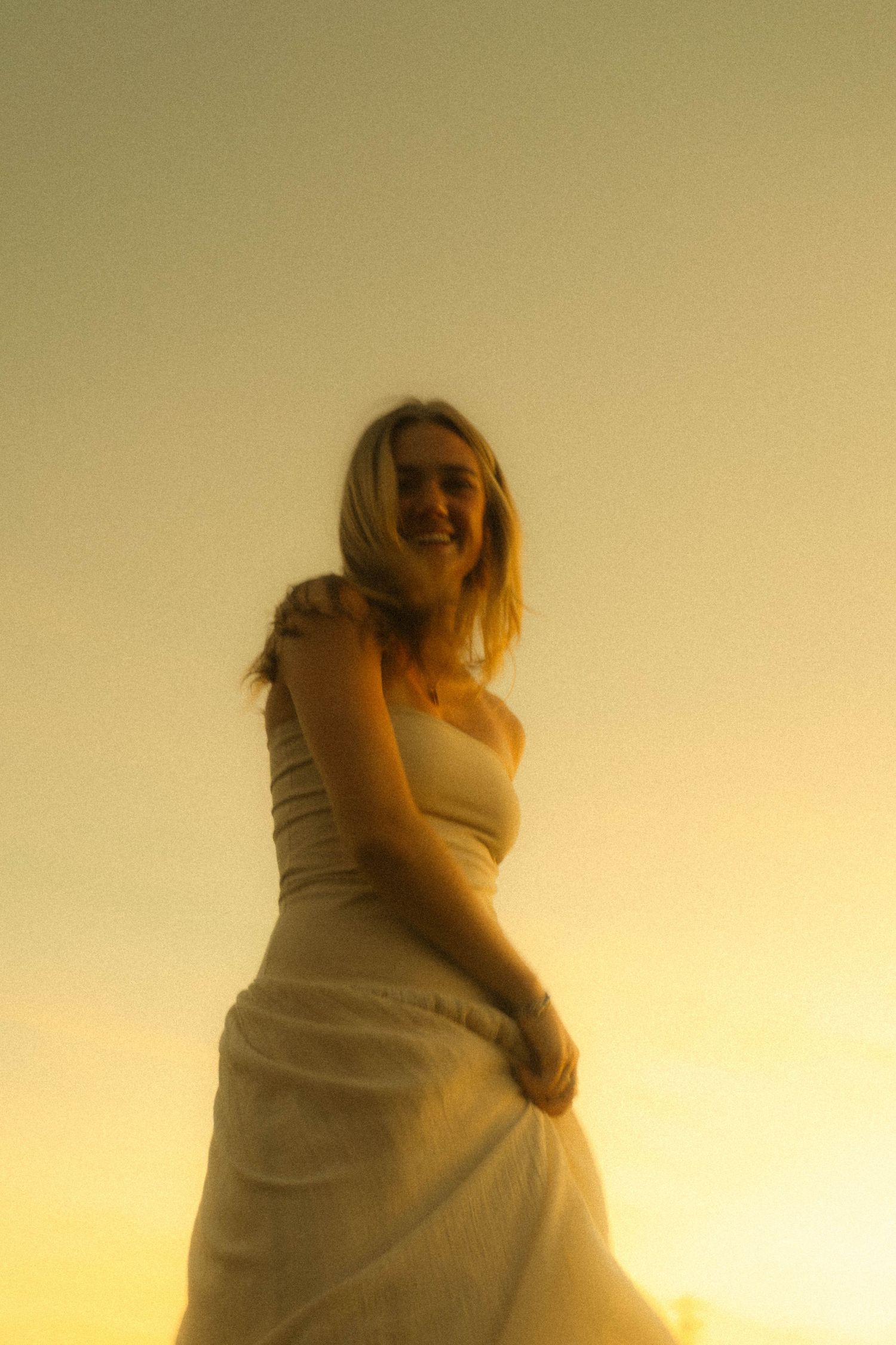 Smiling blonde woman in white dress bathed in warm golden sunset light.