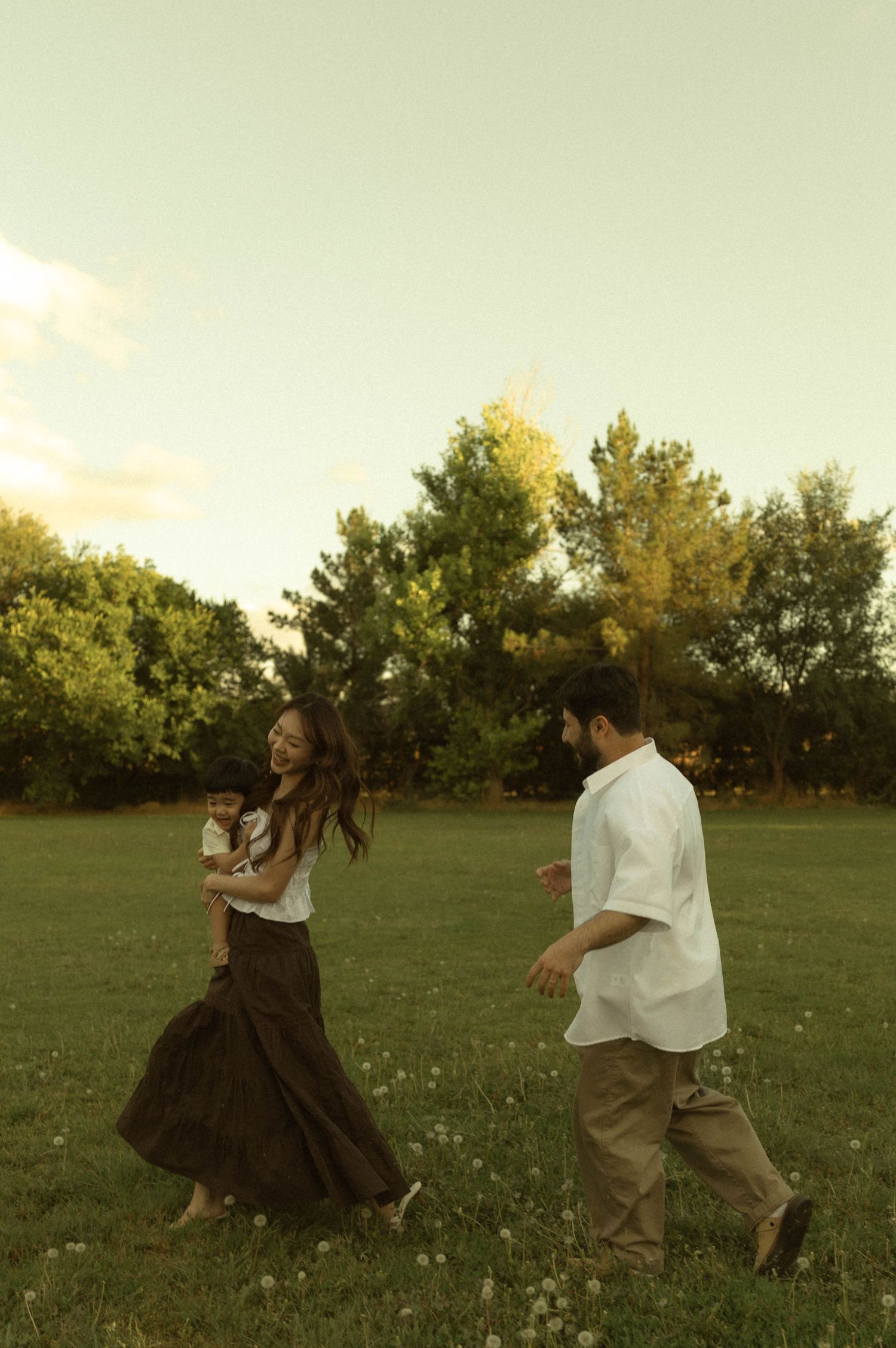A joyful family walks through a lush green meadow at golden hour, woman in dark skirt holding baby.