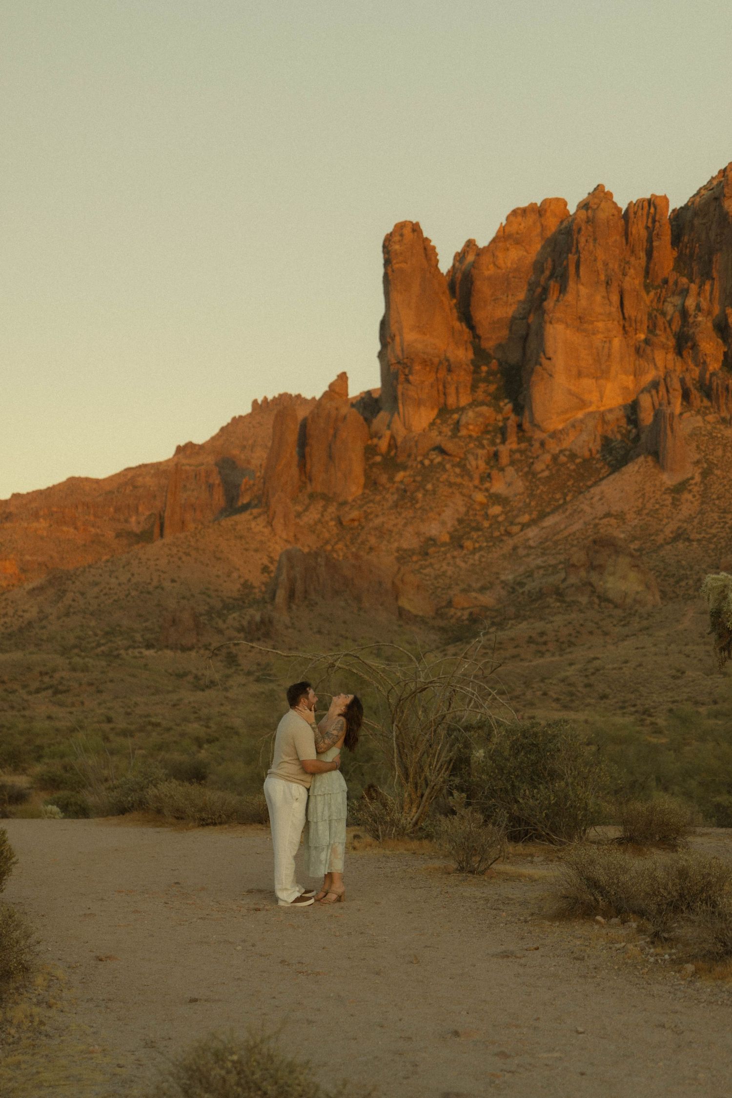Couple embracing at sunset with glowing red rock formations of Superstition Mountains in Arizona desert background.