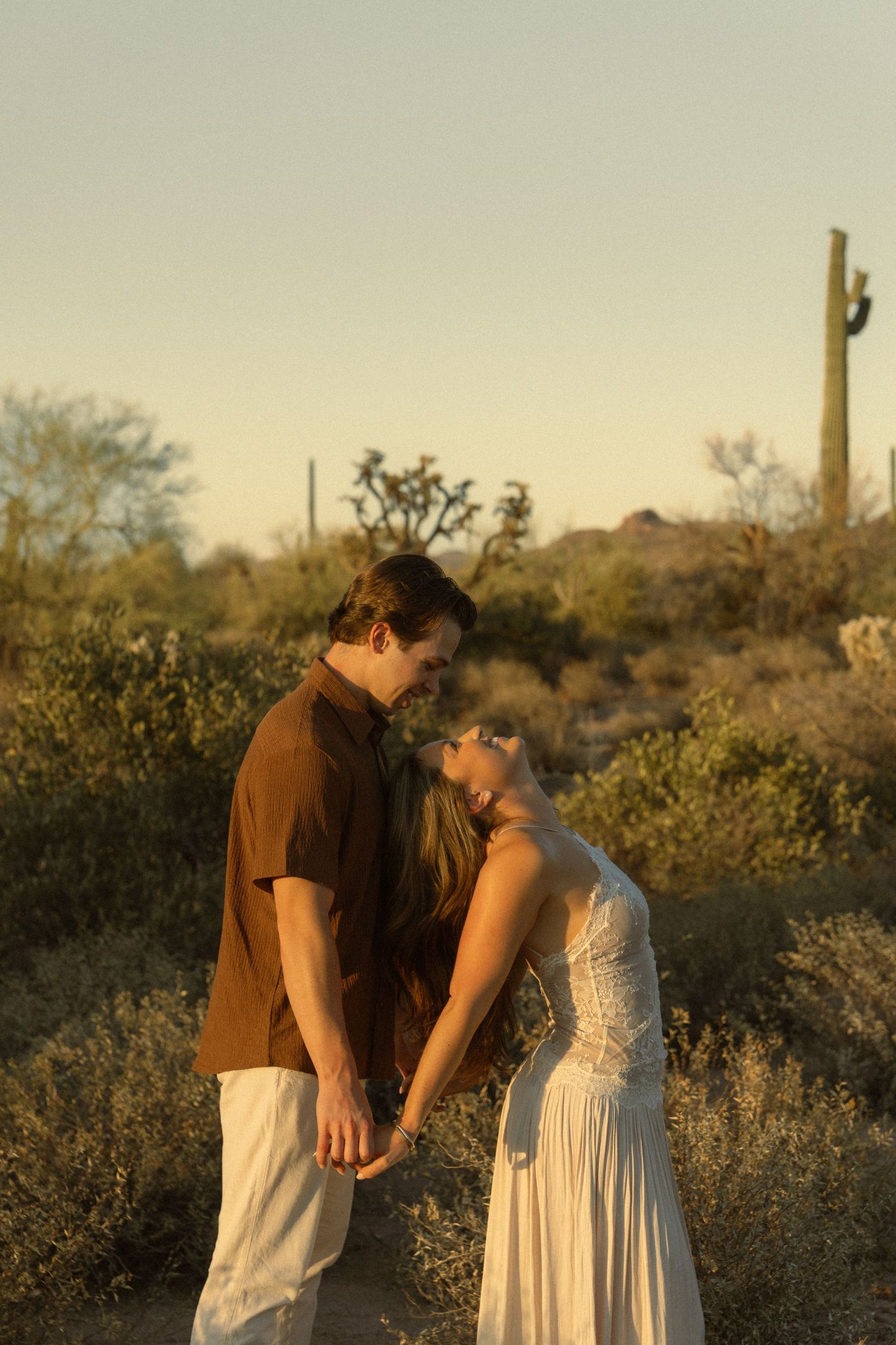 Couple holding hands in a desert landscape at golden hour with saguaro cacti, man in brown shirt and woman in white dress.