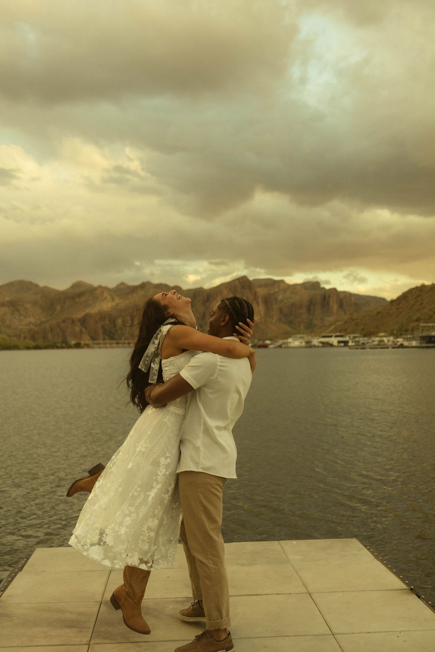 Couple embracing romantically on a waterfront dock with mountain backdrop and dramatic cloudy sunset sky.