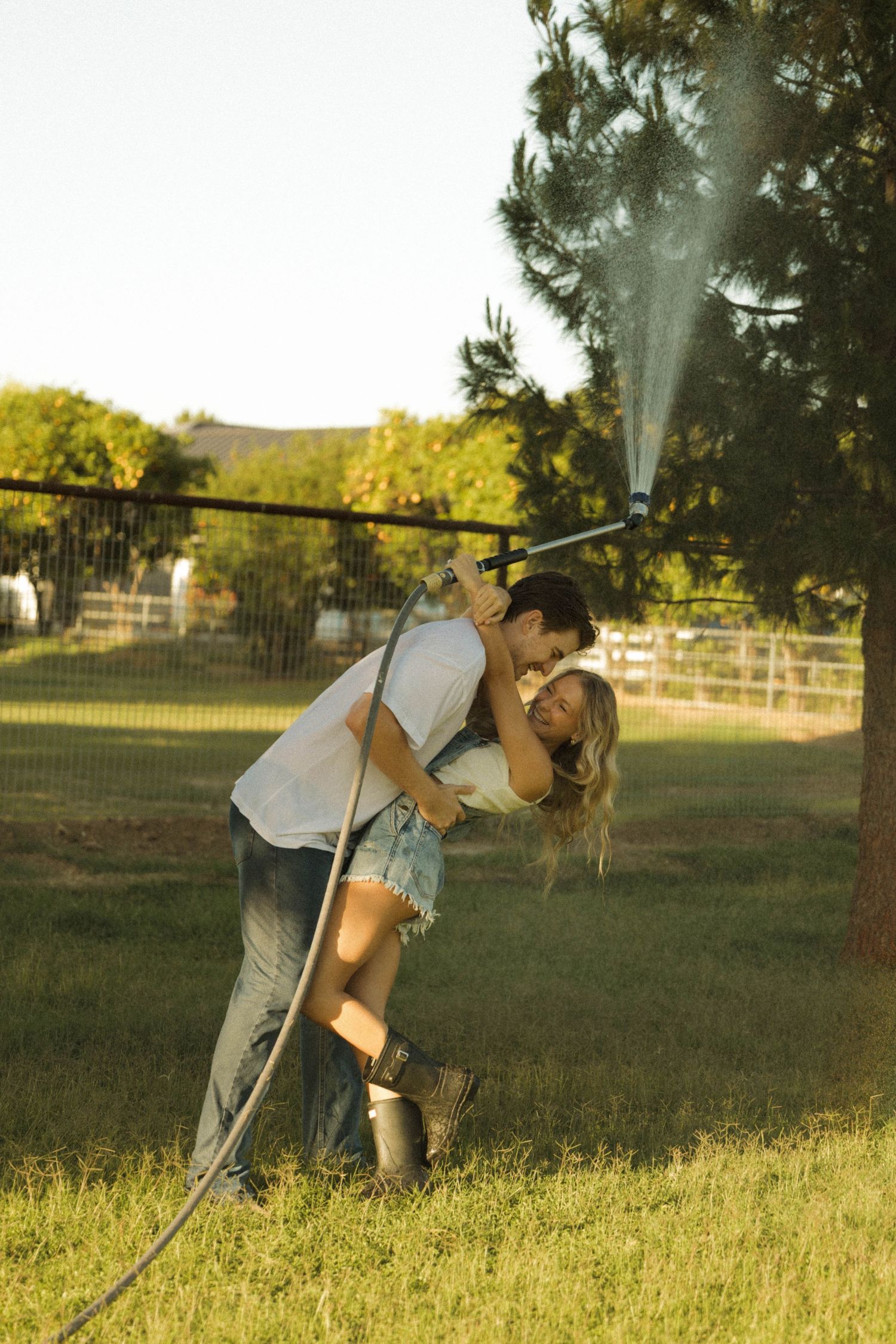 A man dips a woman for a kiss outdoors while holding a spraying garden hose in a green yard.