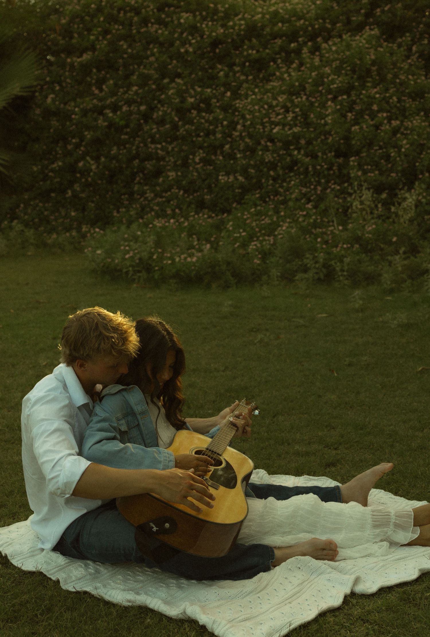 Couple sharing intimate moment on white blanket in lush garden, man strumming acoustic guitar while woman leans close.