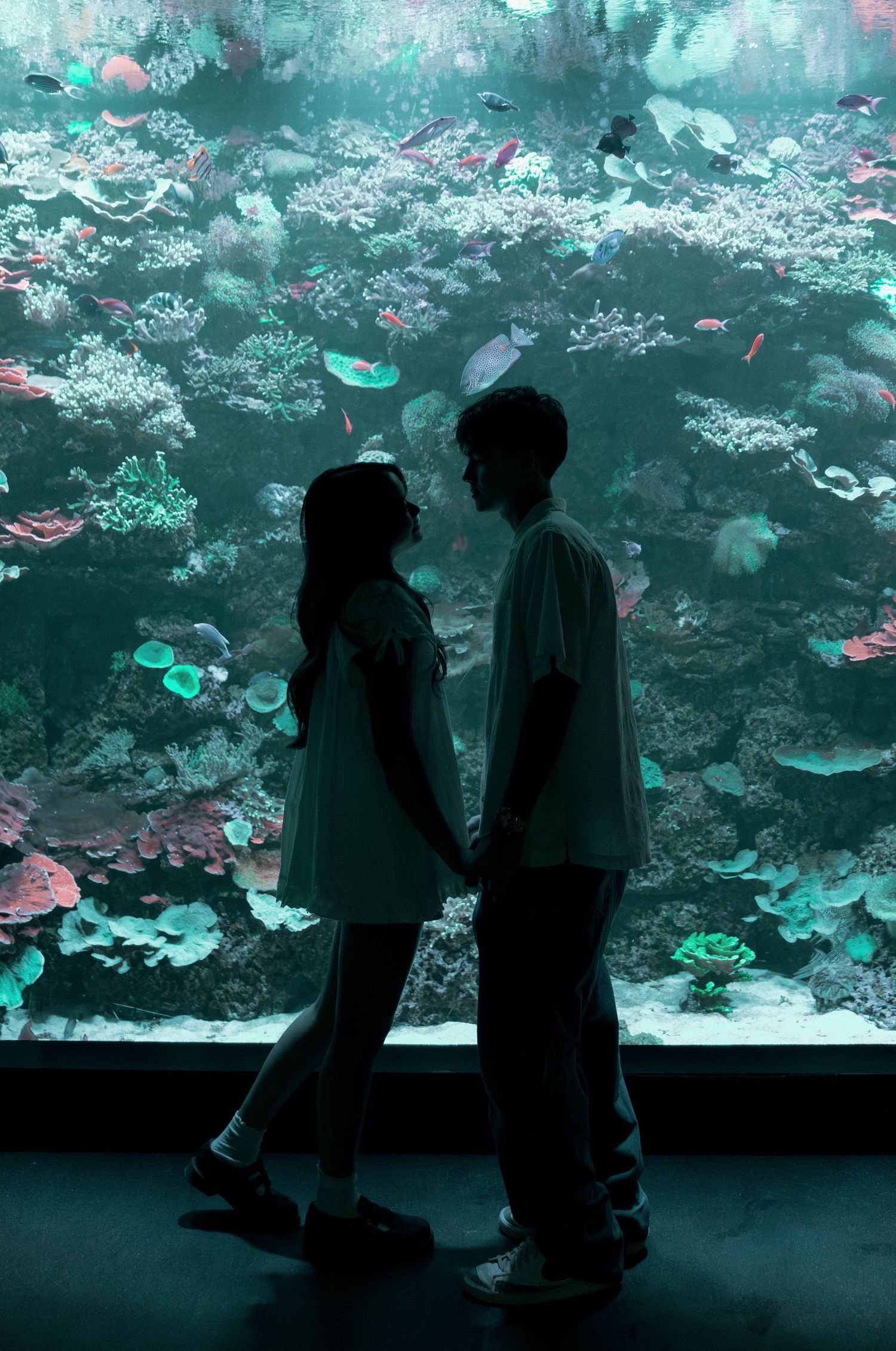 People viewing a large aquarium display with colorful coral and fish in turquoise-tinted lighting.