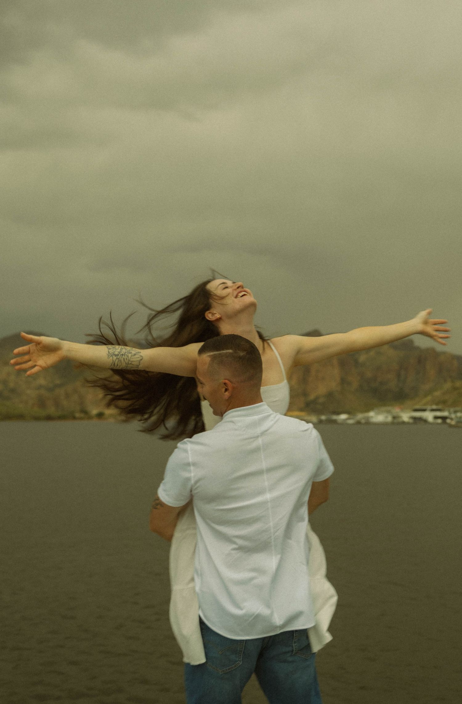 A joyful moment captured as a couple spins by the waterfront with mountains in the background.