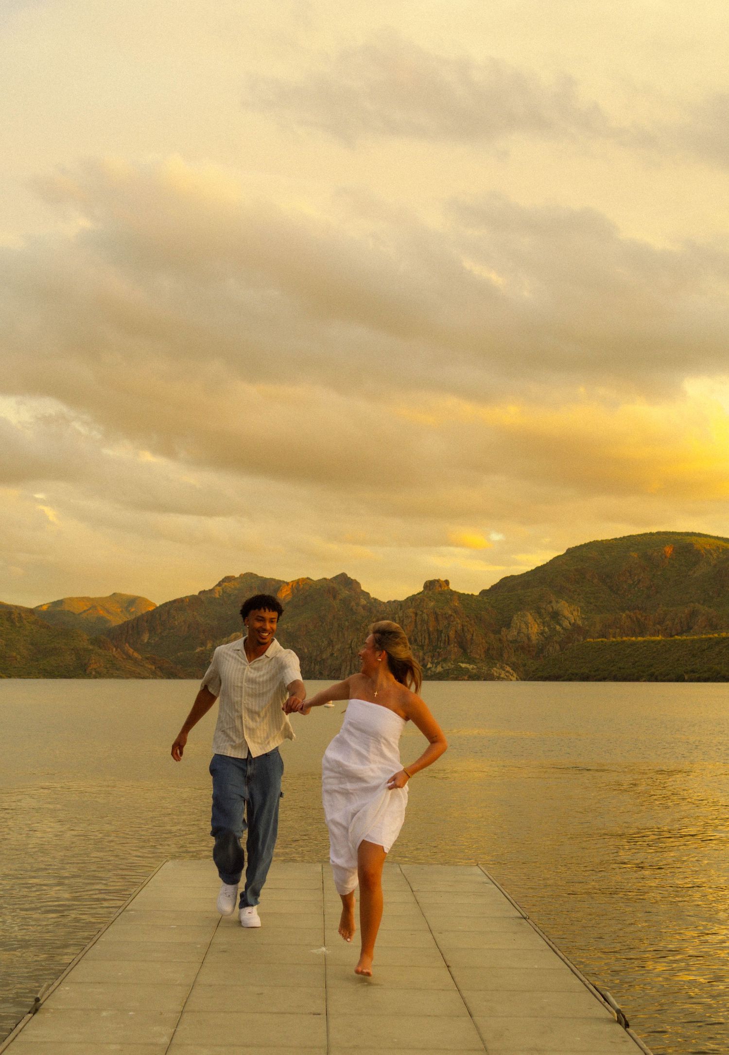 Couple running and playing on a seaside walkway during golden sunset with mountains in the background.