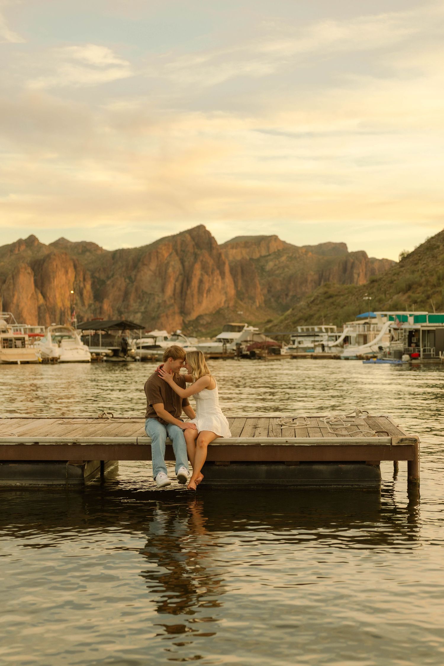 A couple shares a romantic moment on a wooden dock at sunset with mountains and boats in the background.