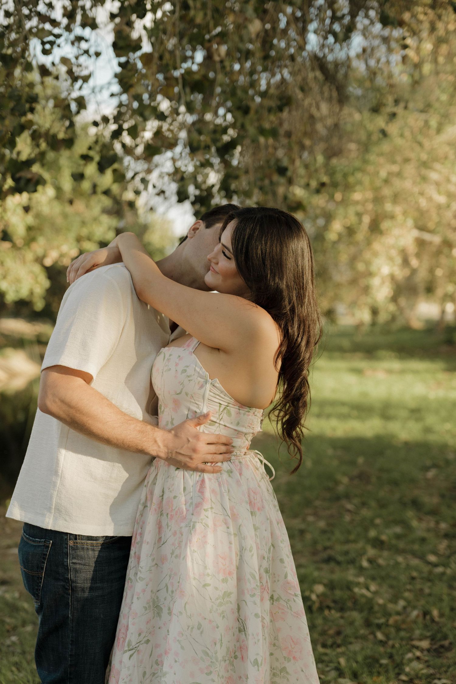 A romantic couple embraces outdoors in a garden setting, with her wearing a floral dress against golden sunlit trees.