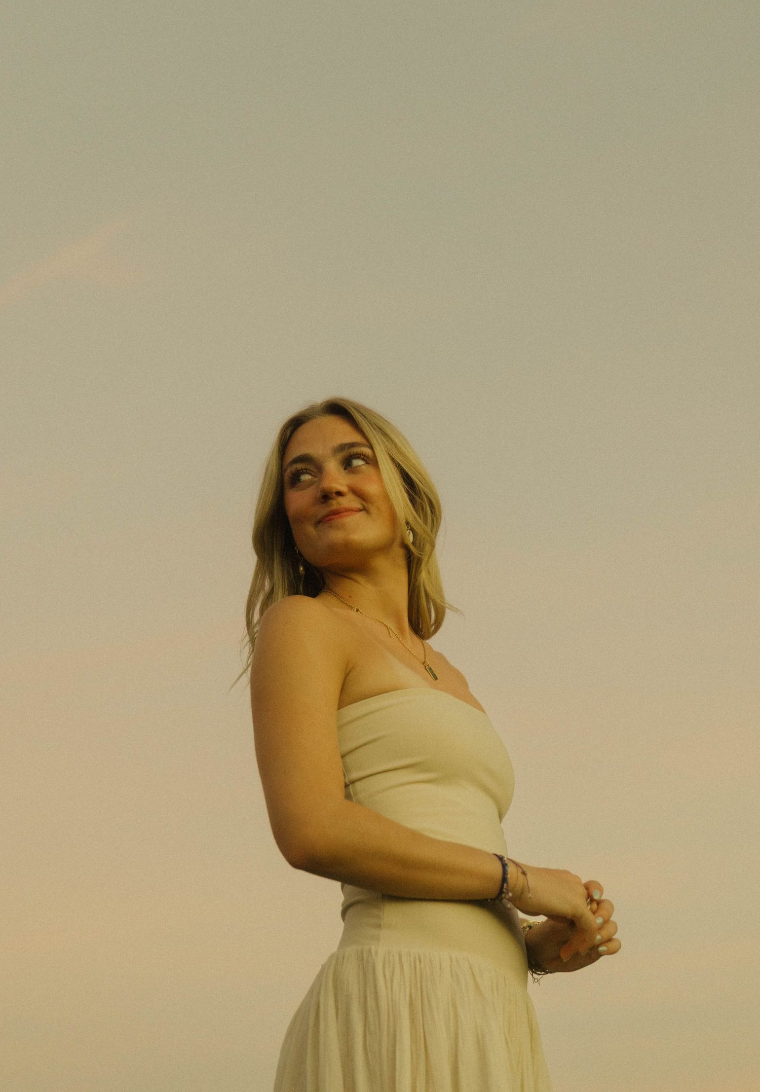 Young blonde woman in white strapless dress gazing upward against a soft pastel sky.
