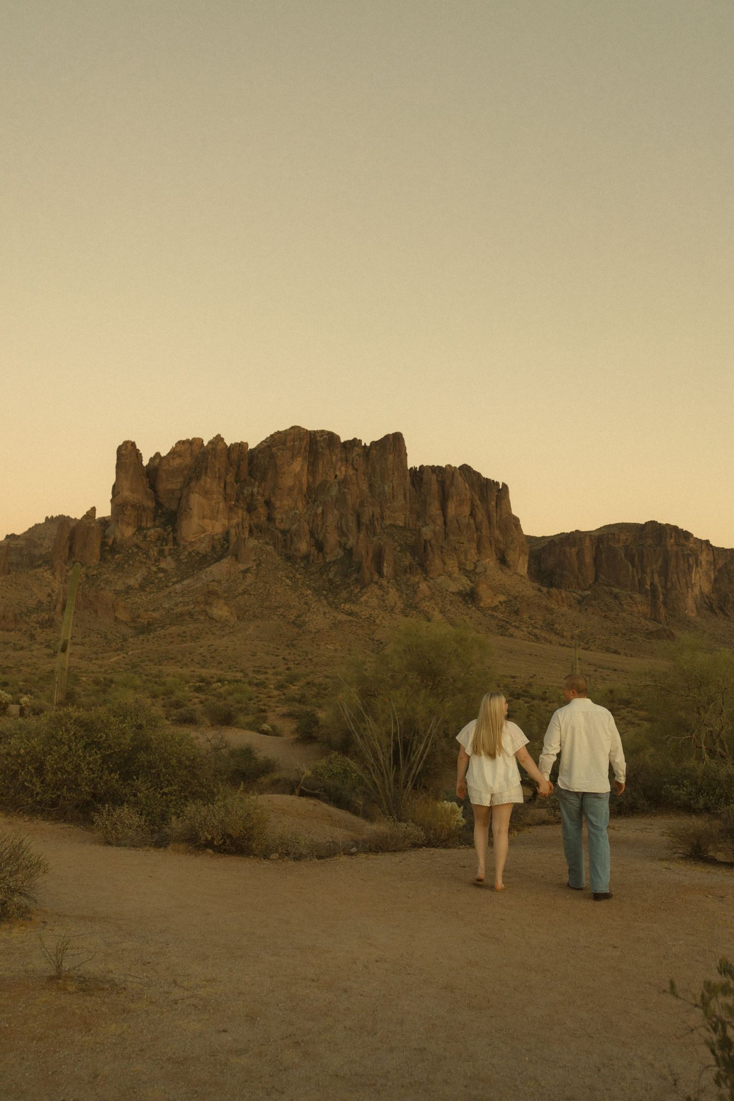 Couple holding hands walking toward Superstition Mountains in Arizona desert at golden hour sunset.