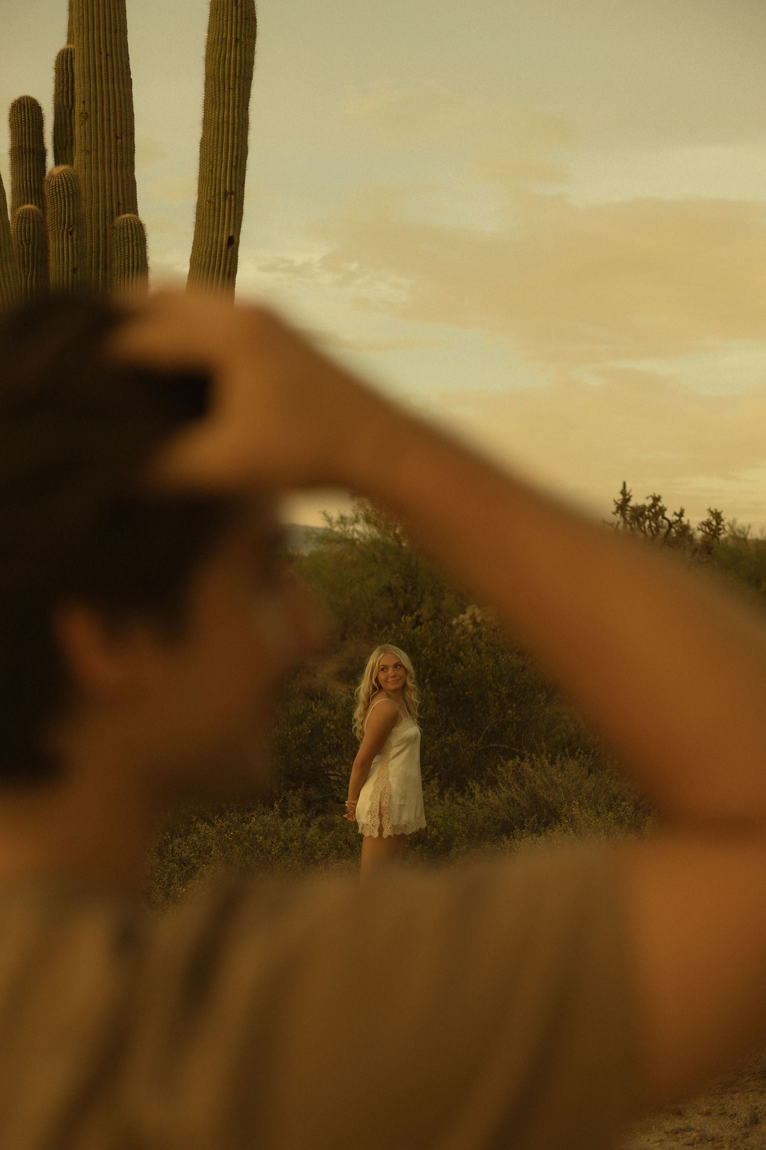 Man photographing blonde woman standing among saguaro cacti in desert at sunset.