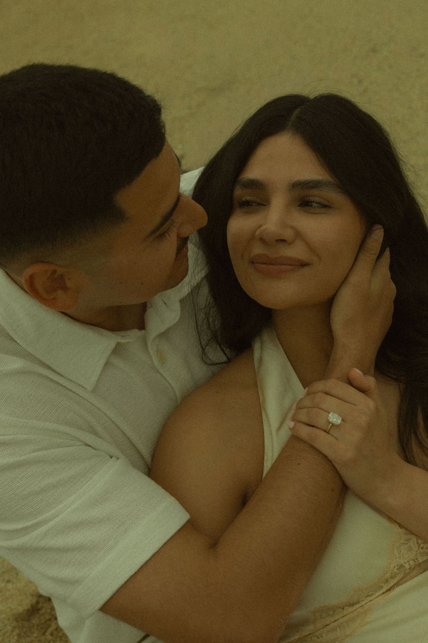 A couple shares a romantic moment, the woman wearing an engagement ring while her partner tenderly holds her face close.