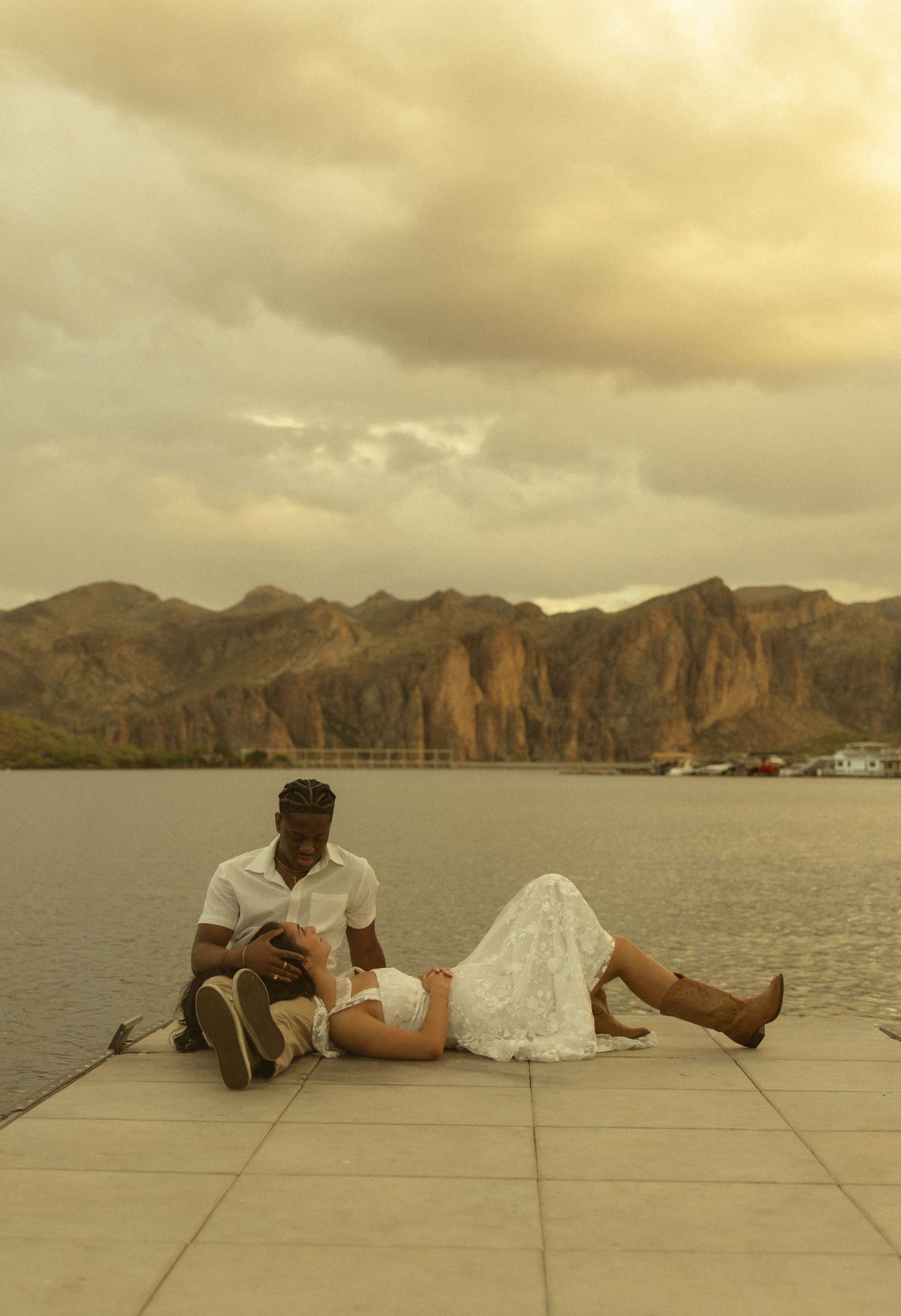Couple relaxing on a lakeside pier with breathtaking mountain landscape under a dramatic cloudy sky.