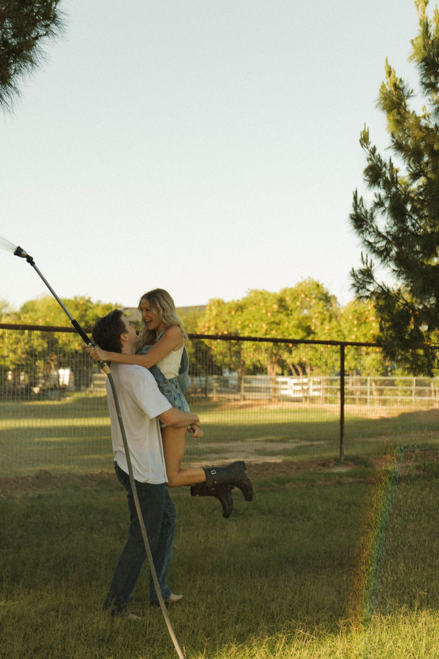 Man lifting woman in a joyful embrace while holding a garden hose in a sunny backyard.