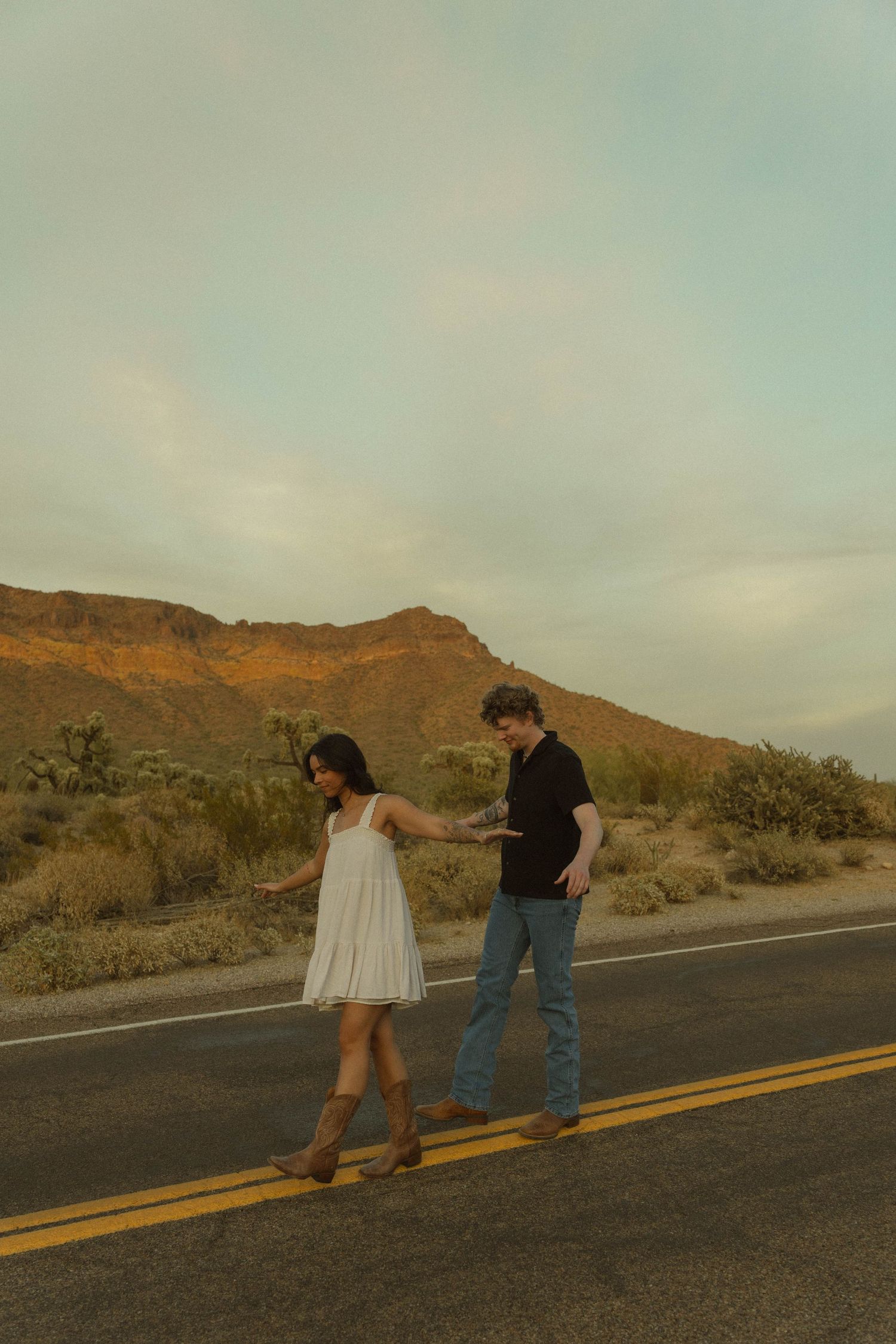 Couple holding hands walking on a desert highway at sunset with mountains in the background.