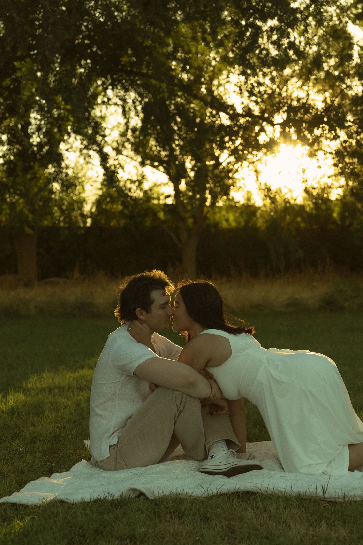 Romantic couple sharing a kiss on a blanket in a lush green park at golden hour sunset.