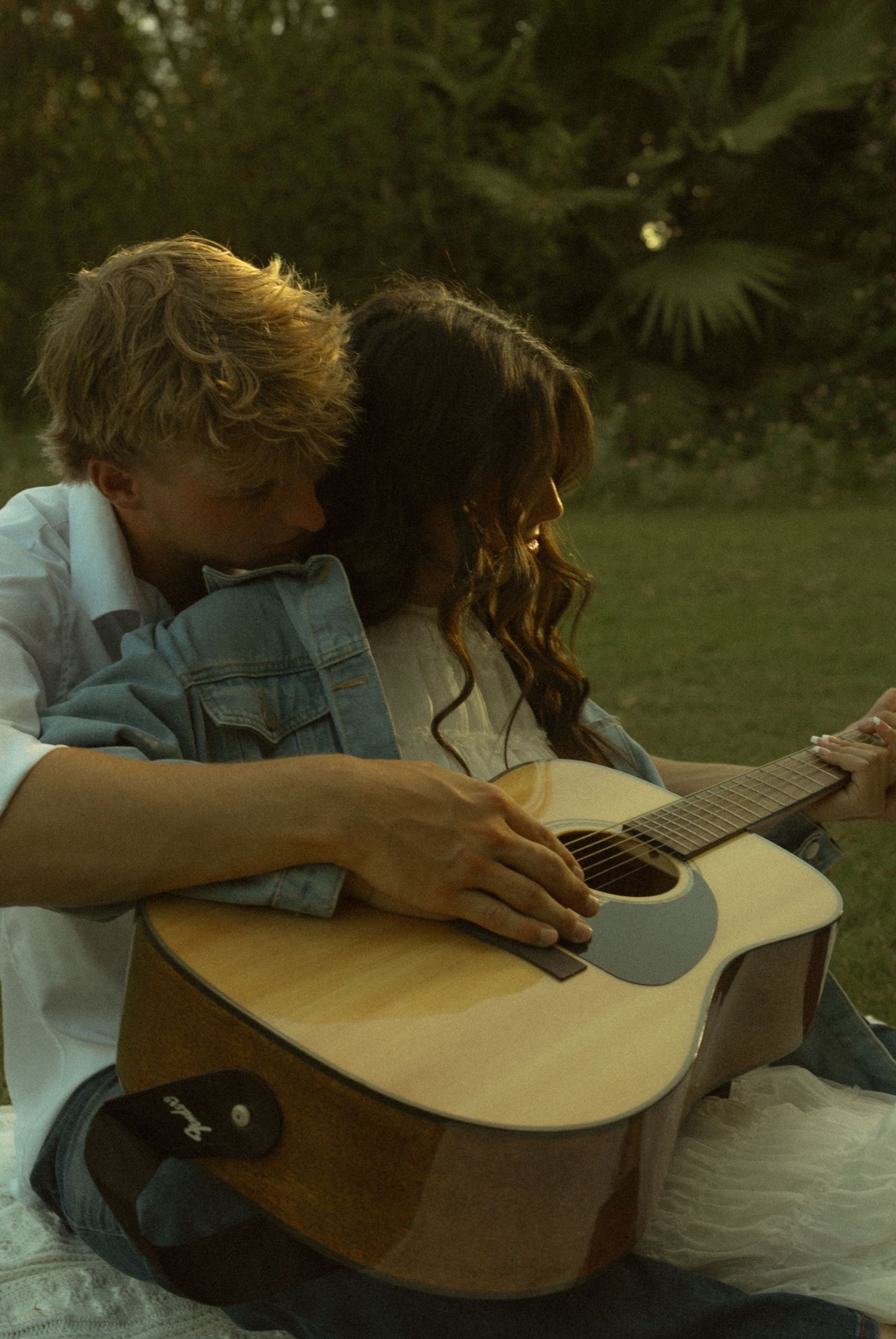A couple sits outdoors sharing an acoustic guitar, with the man leaning in to kiss the woman's cheek.