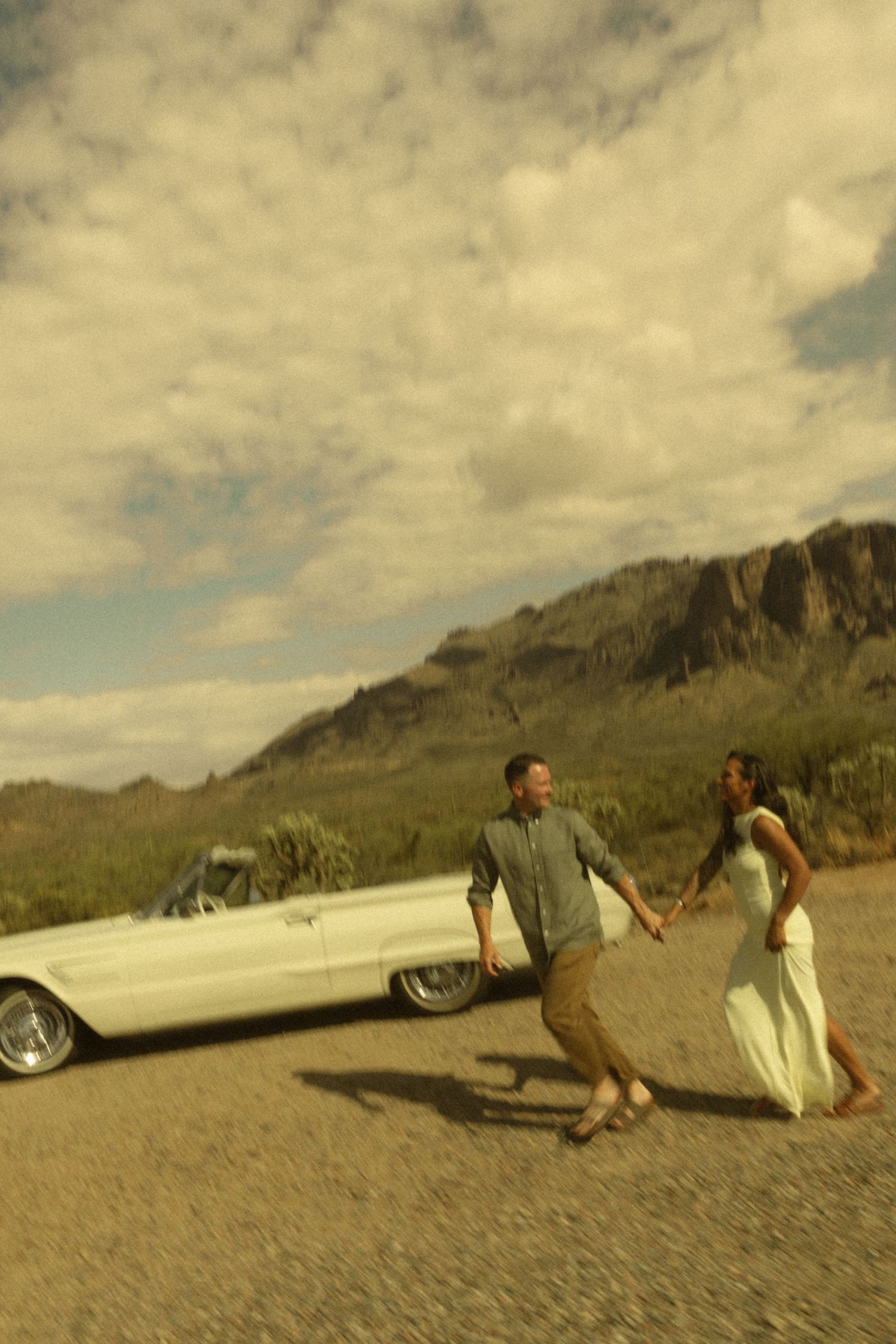 Newlyweds holding hands walking beside a classic white convertible with rocky mountains in background.
