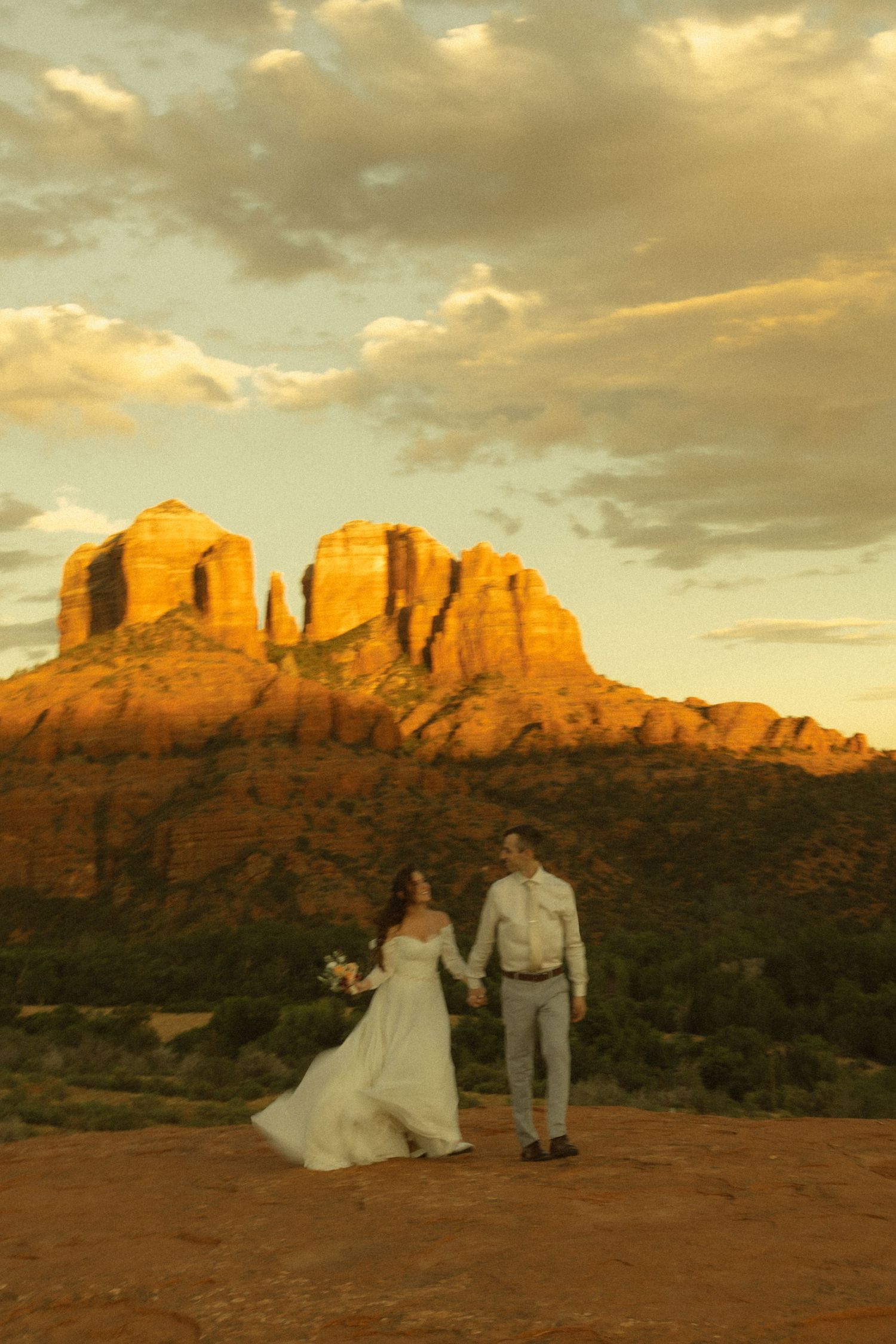 Bride and groom hold hands at sunset with glowing red Cathedral Rock formations in Sedona, Arizona behind them.