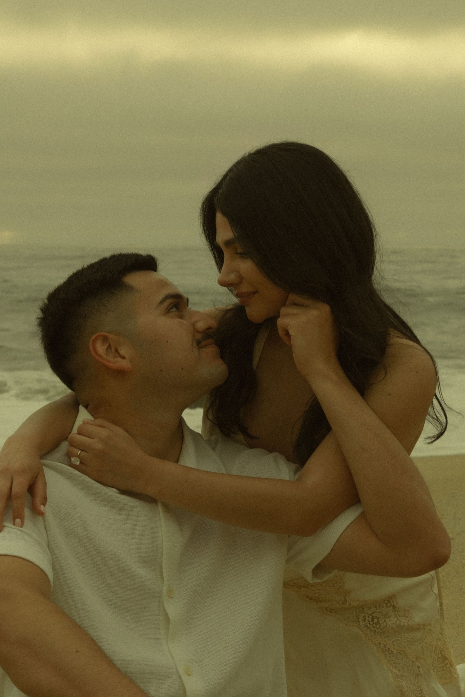 A romantic couple embraces and smiles at each other on a cloudy beach, the woman wearing a ring.