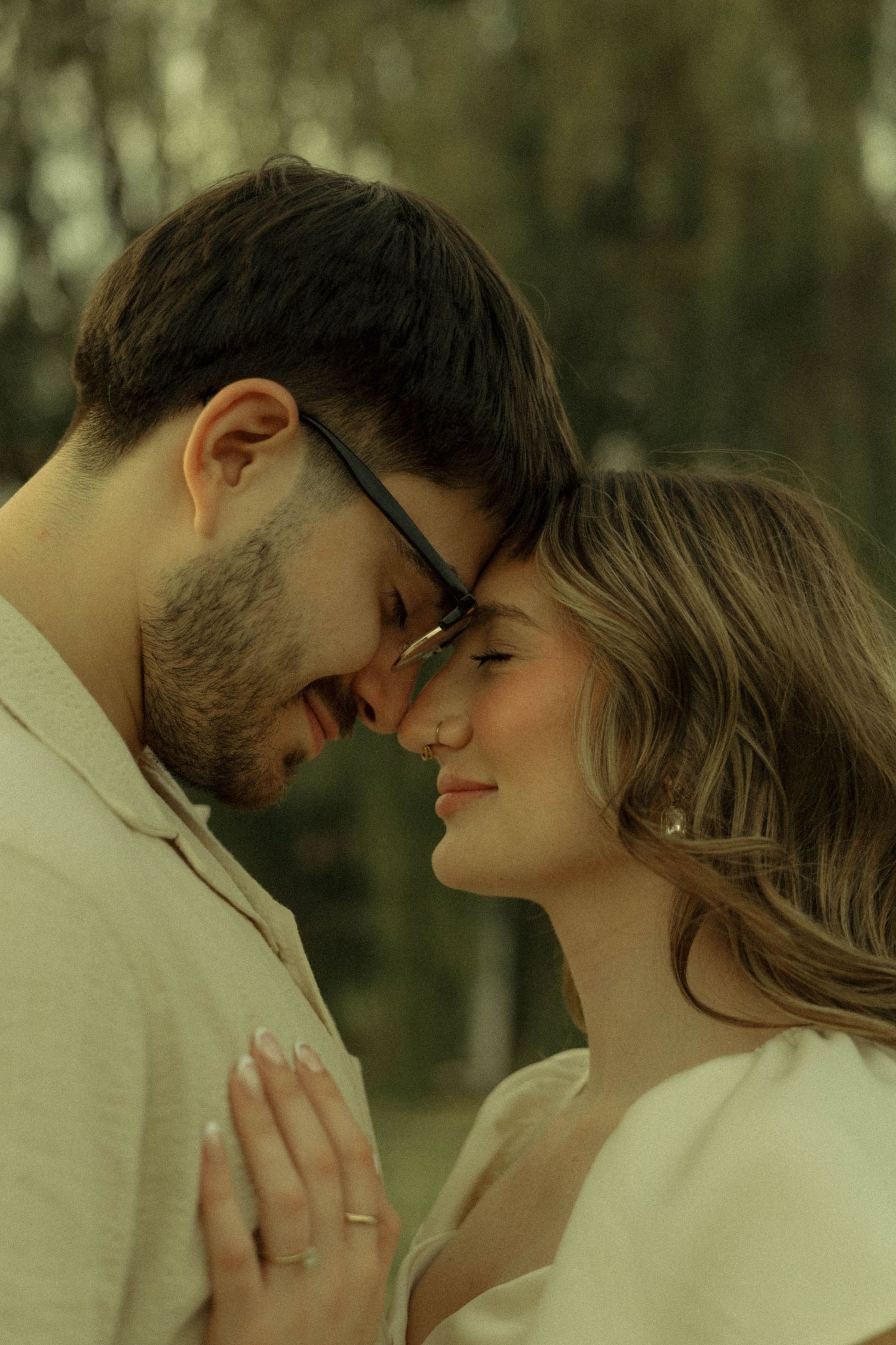 Romantic couple touching foreheads tenderly in a park, man wearing glasses, woman smiling softly, sharing an intimate moment.