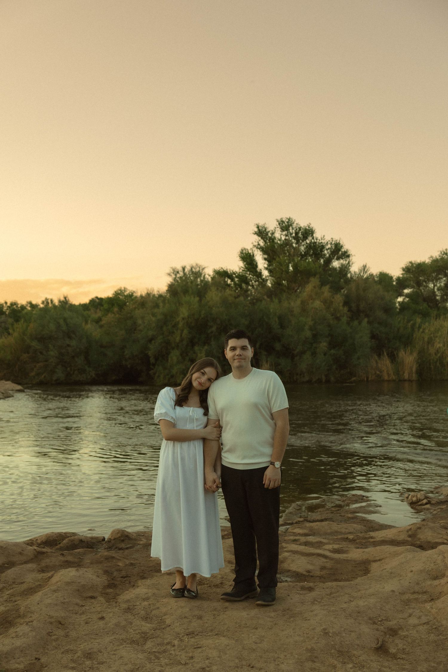 Couple posing together by a peaceful river with trees glowing in sunset light.
