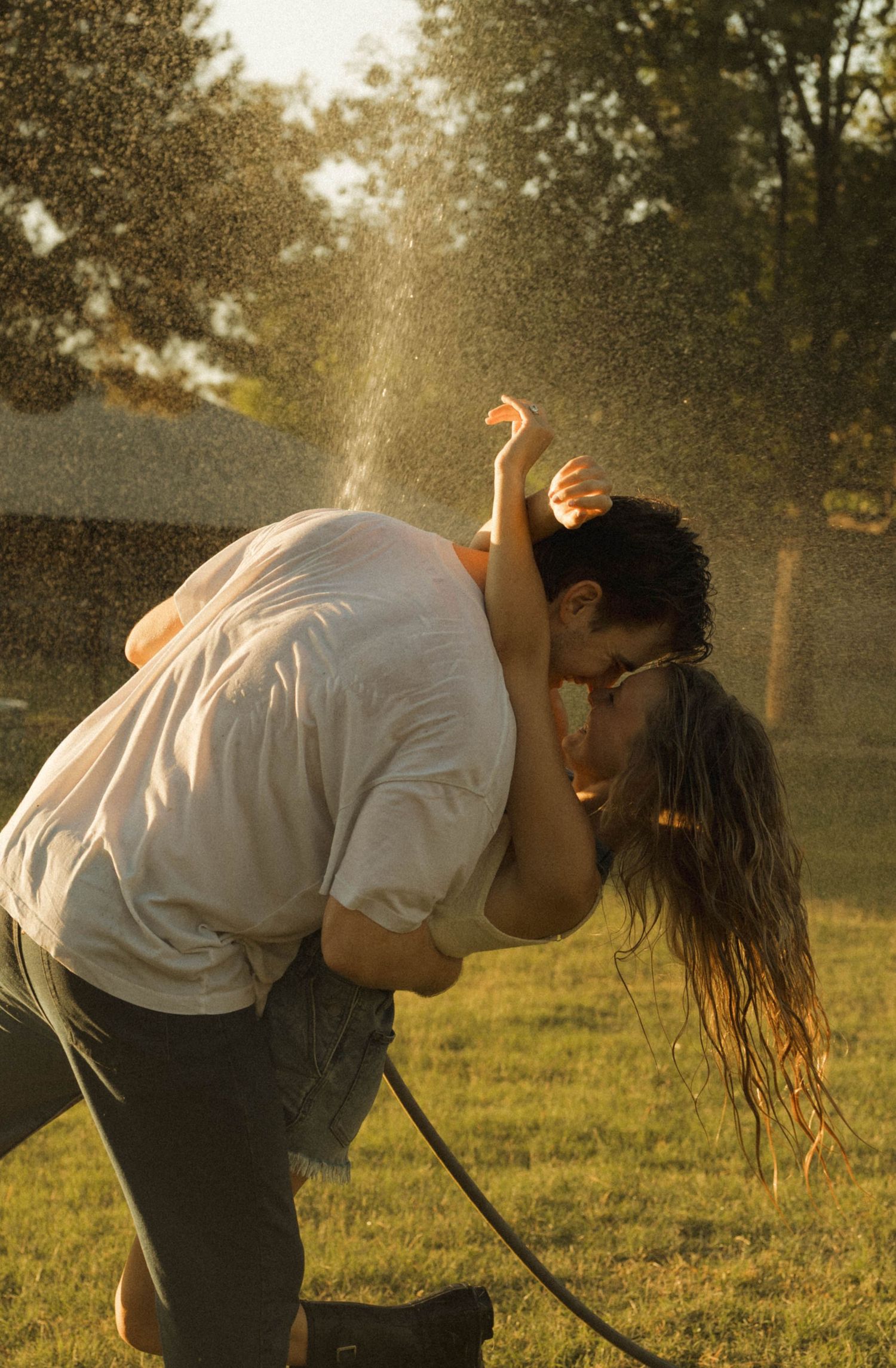 Couple kissing and playing under a garden hose sprinkler in a sunny backyard on a warm summer day.