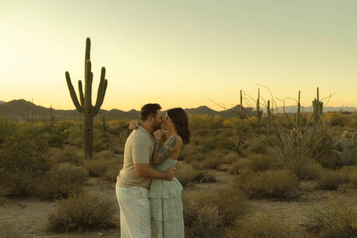 Couple sharing a romantic moment at sunset in the Sonoran Desert with saguaro cacti and mountains in the background.