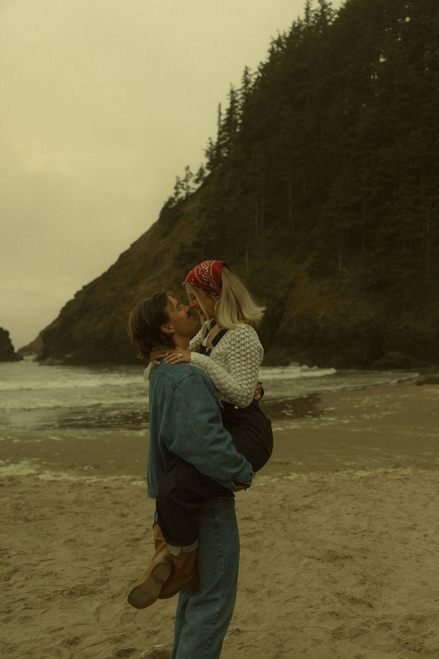 Couple embracing and kissing on a misty beach with rocky cliffs and evergreen trees in the background.