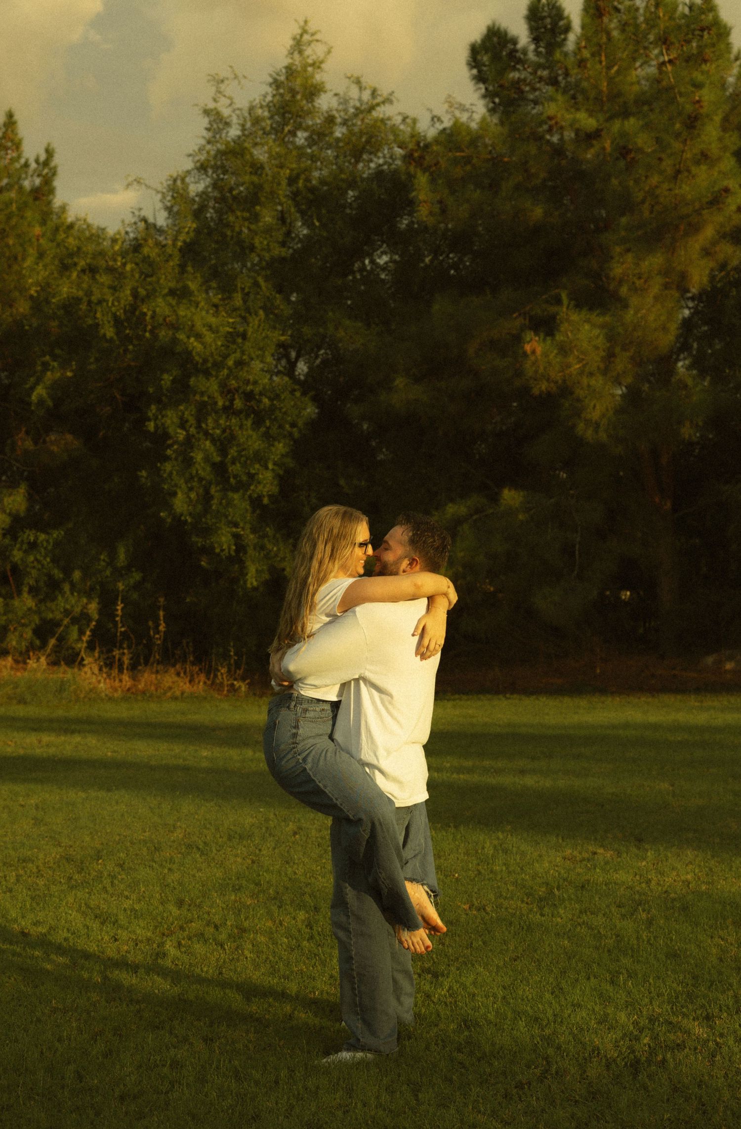 A couple embraces romantically in a sunlit green park, the man lifting the woman as they share a tender moment.