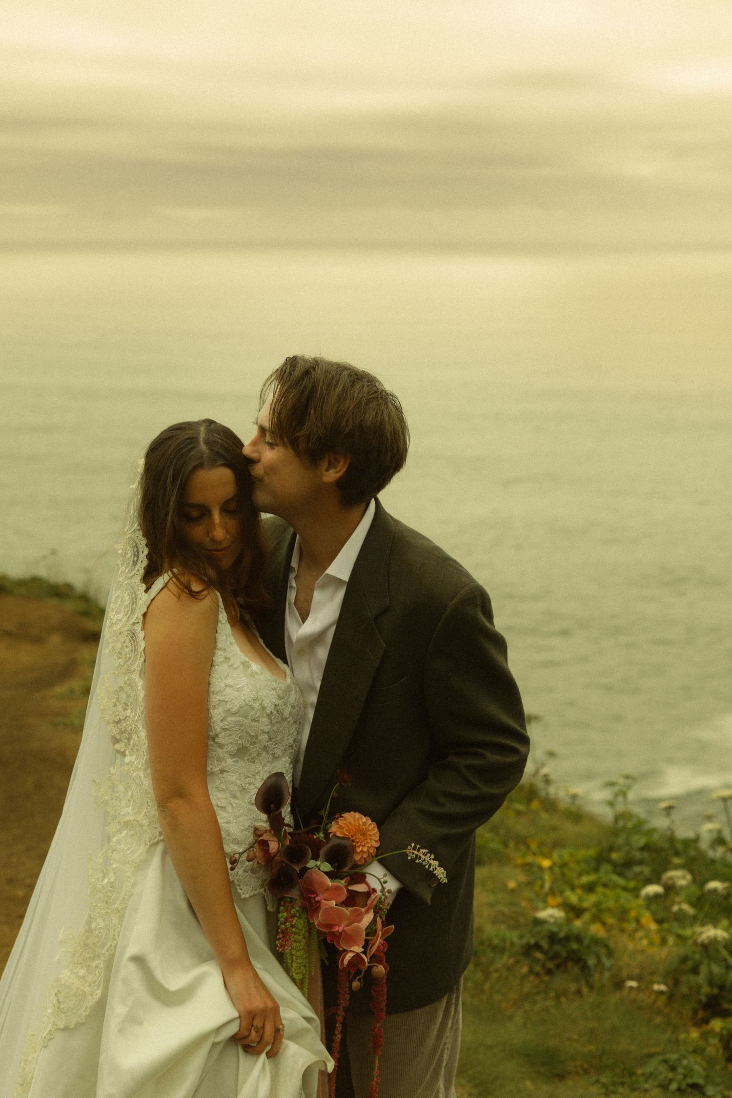 Bride and groom share a romantic moment on a coastal cliff at sunset, with ocean views behind them.