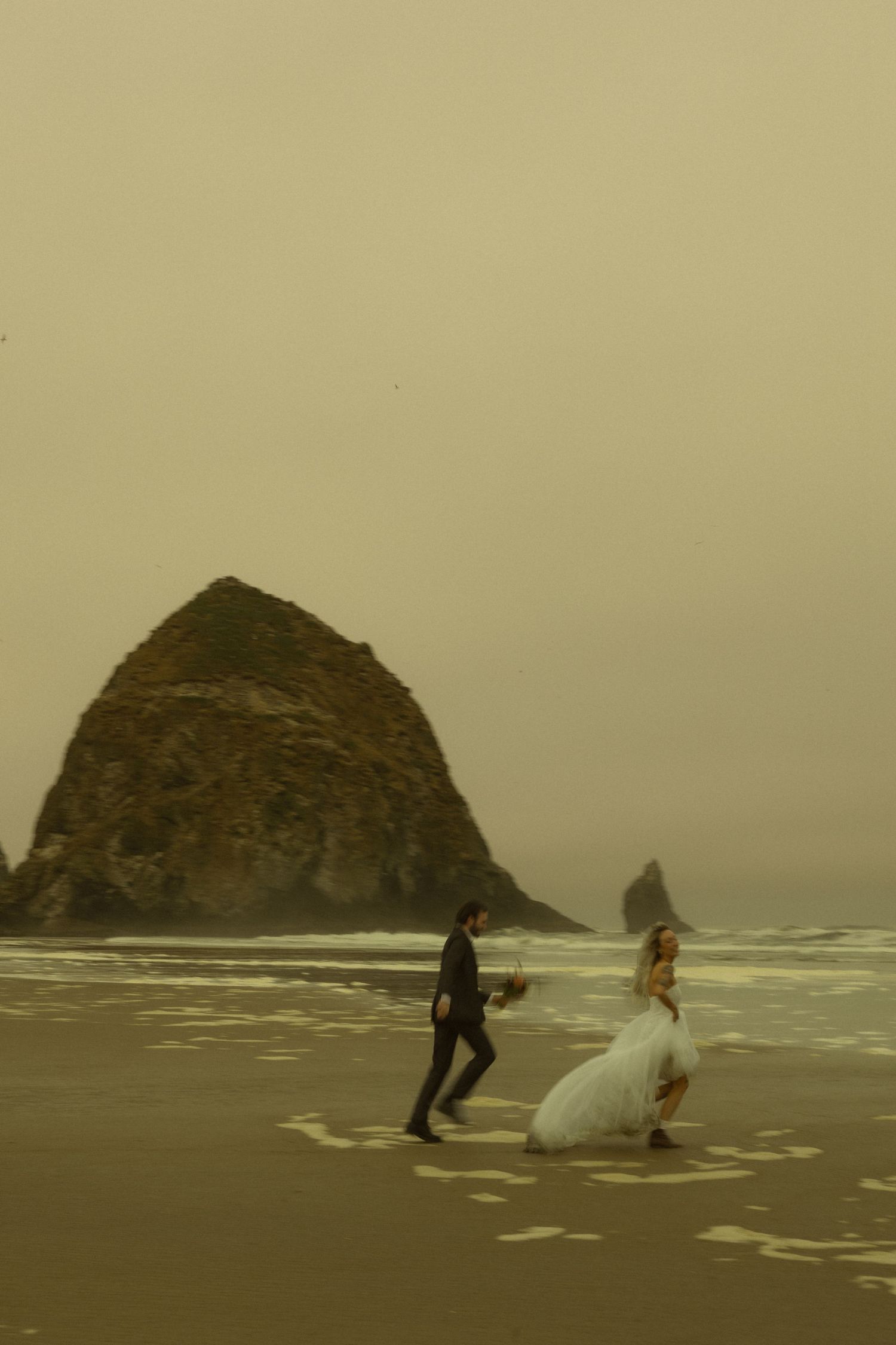 Bride and groom walk on misty beach near Haystack Rock at Cannon Beach, Oregon wedding elopement.