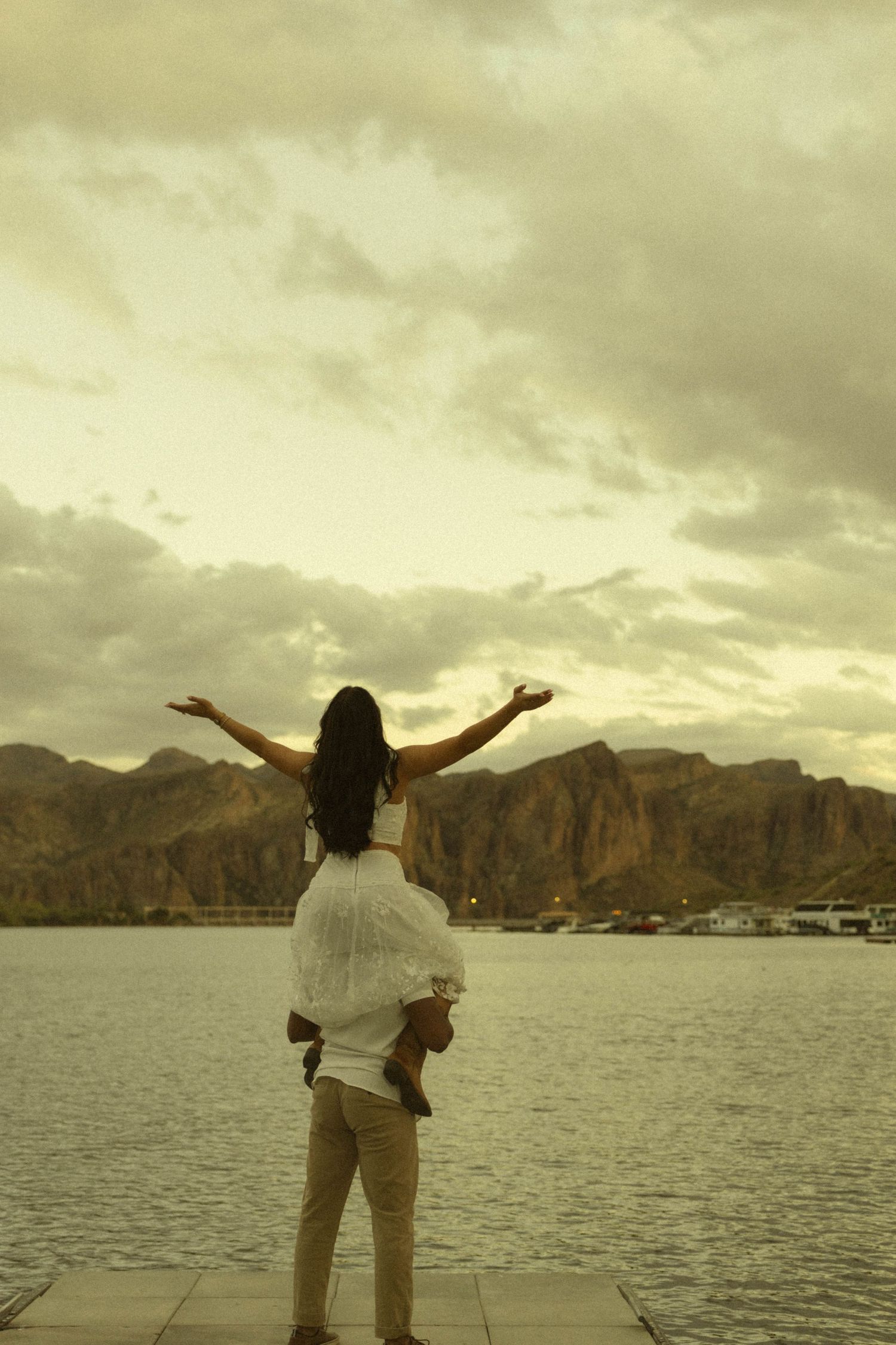 A man carries a woman on his back as she spreads her arms wide facing a scenic mountain lake at sunset.
