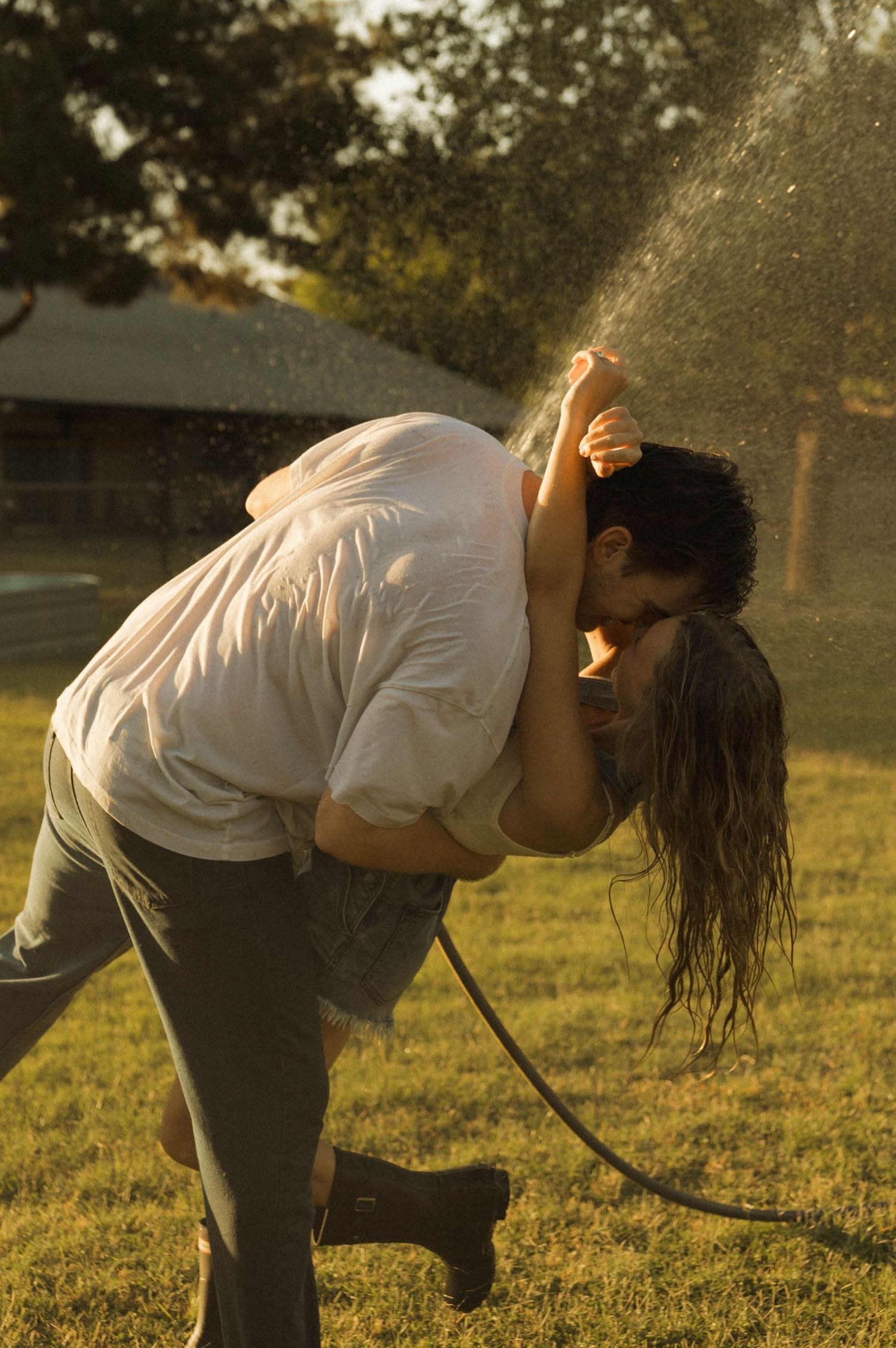 Couple sharing a passionate kiss while getting soaked by a garden hose in a sunlit backyard.