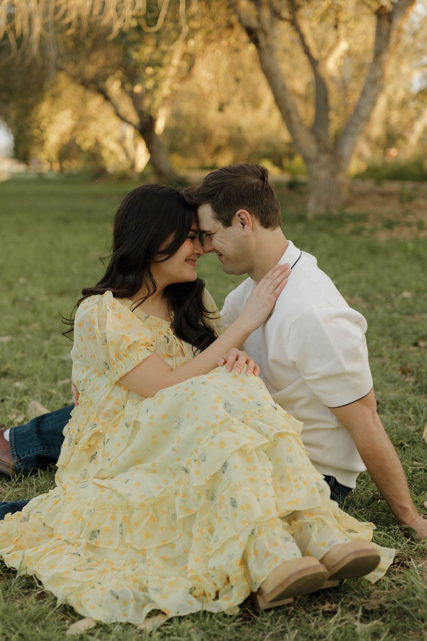 A romantic scene unfolds as a couple in casual attire embraces on grassy ground during sunset.