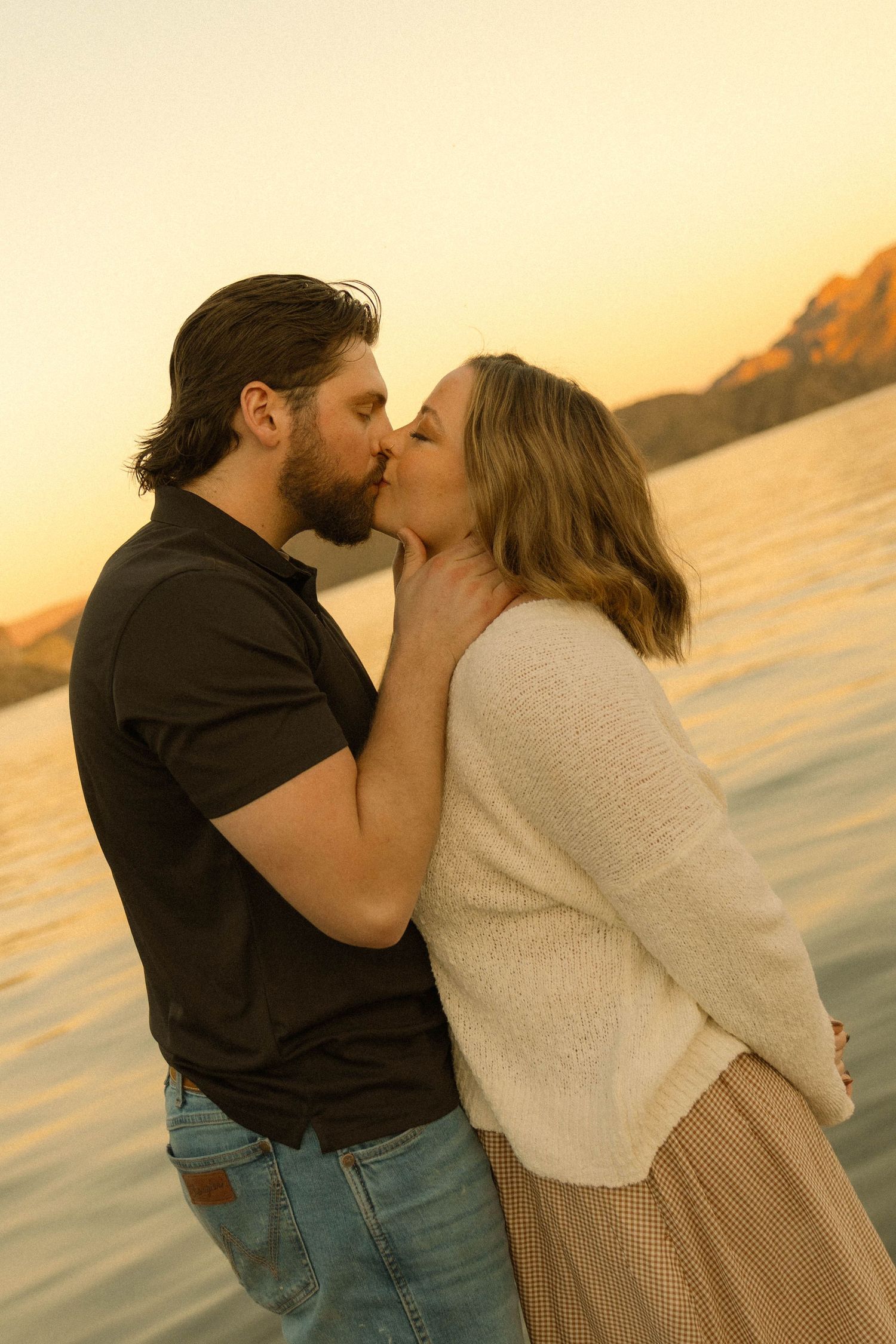 Tender kiss shared between couple against backdrop of sunset over water.