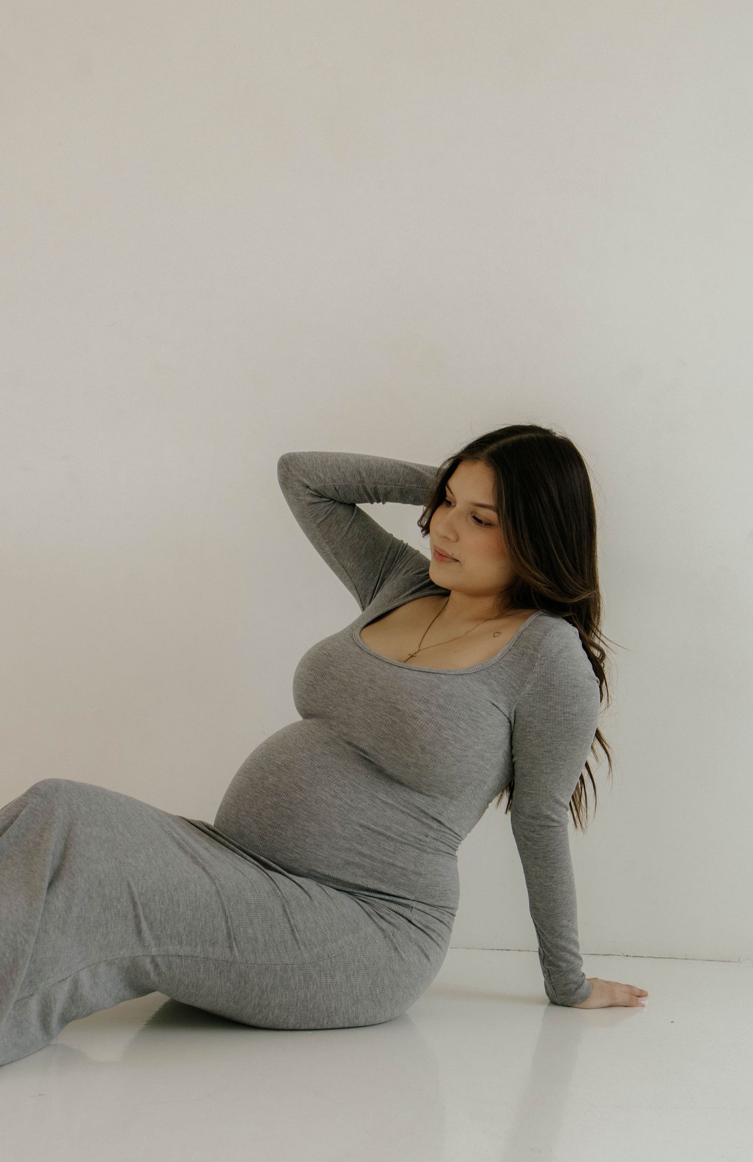 A pregnant woman in a fitted grey dress reclines on a white floor.