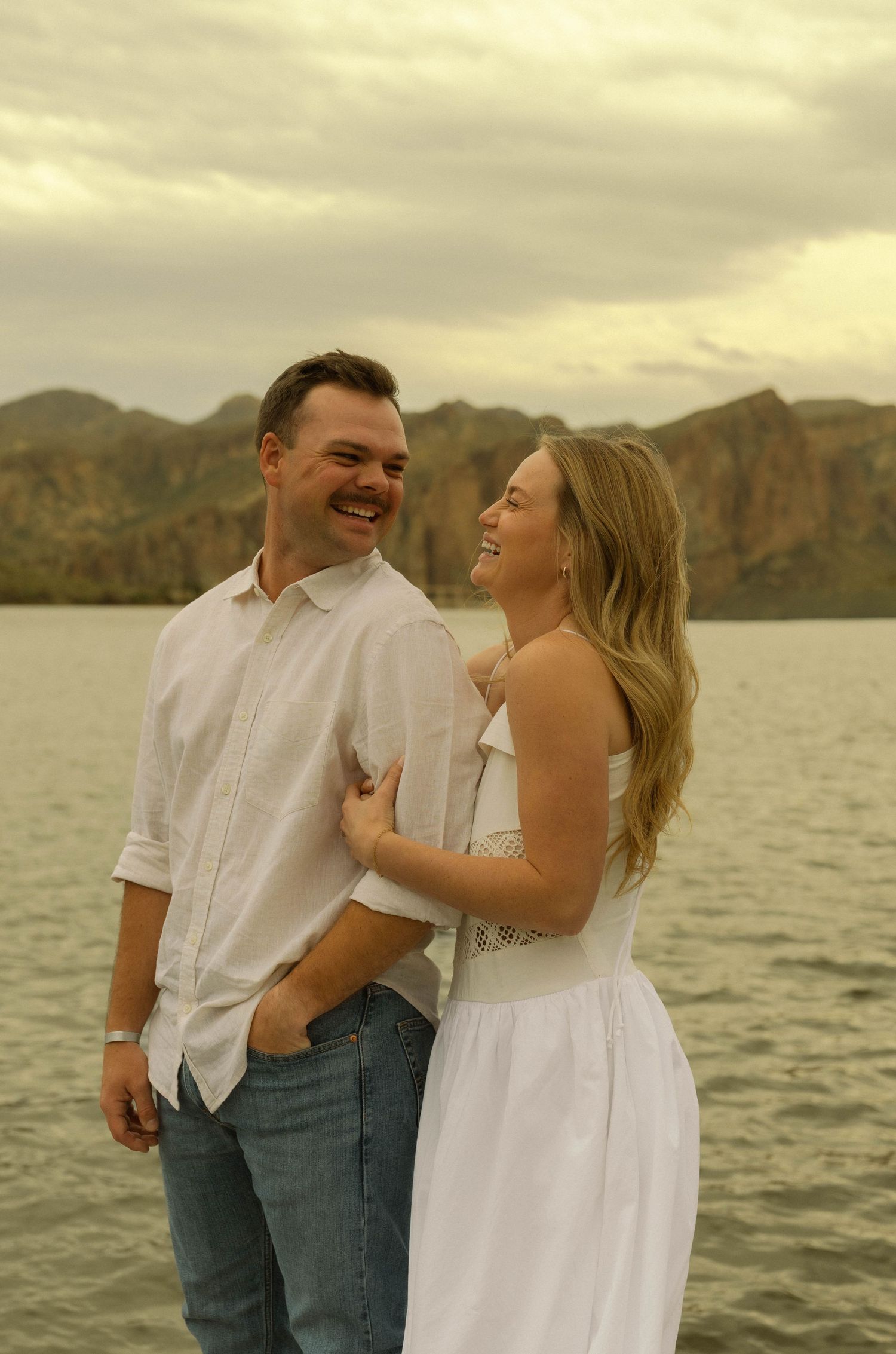 A couple in white clothing shares a joyful moment together by a lake with mountains in the background.