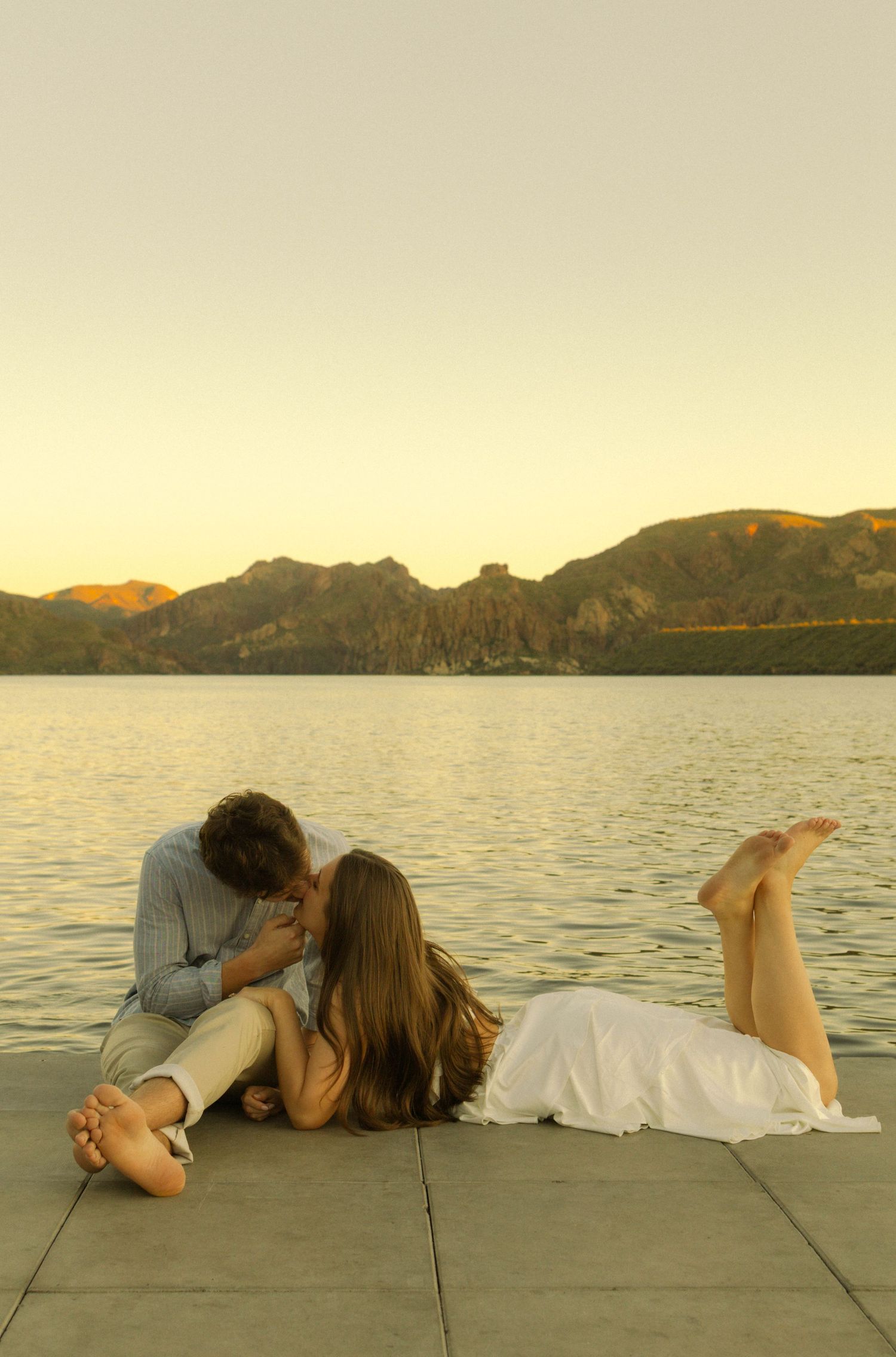 A couple lying together on a dock during golden hour with mountains and water in the distance.