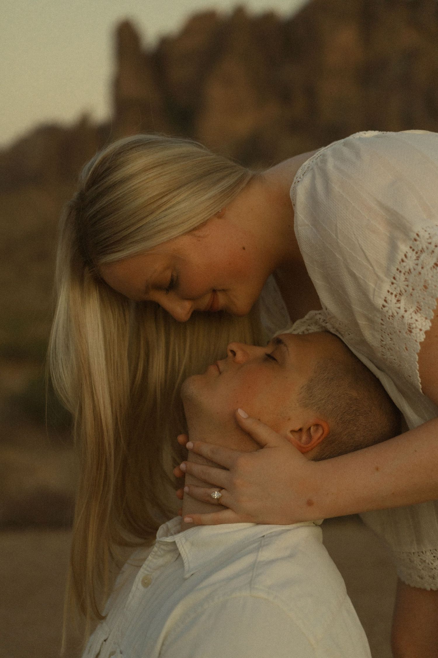 A couple shares a tender romantic moment outdoors, with the woman leaning down to kiss the man against a rocky desert backdrop.