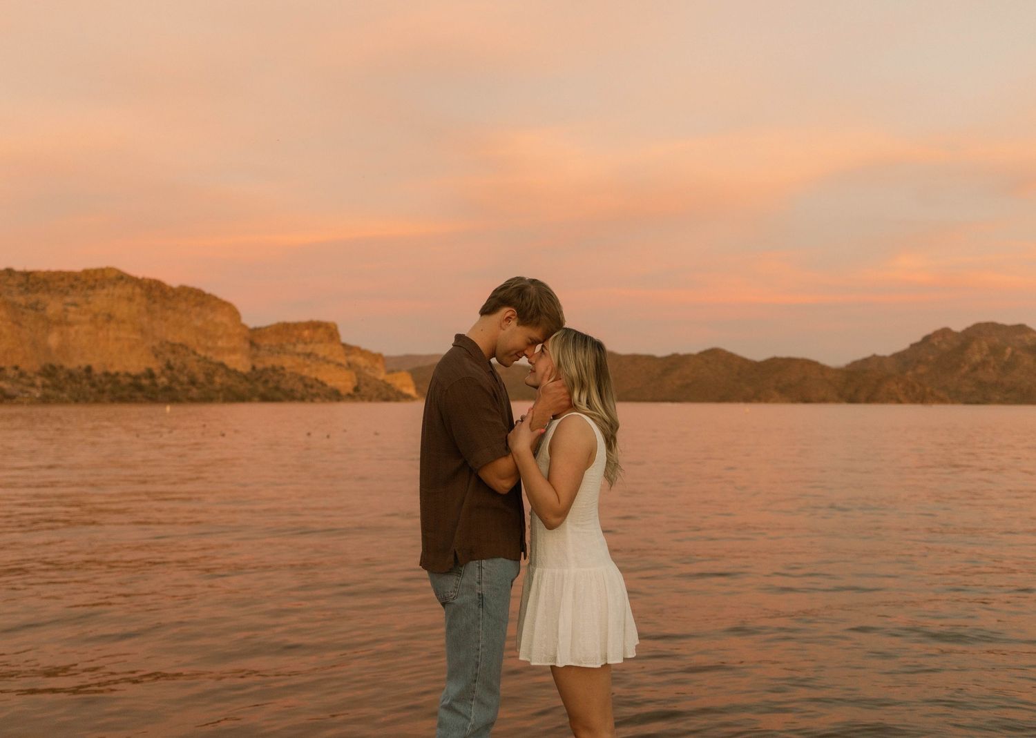Couple sharing a romantic moment by a lake at golden hour with a stunning pink and orange sunset sky.