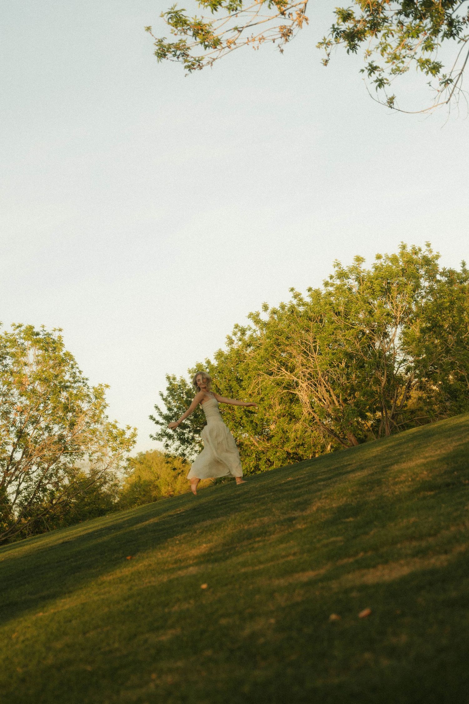 A bride in a white gown walks gracefully across a lush green hillside surrounded by trees in golden hour light.