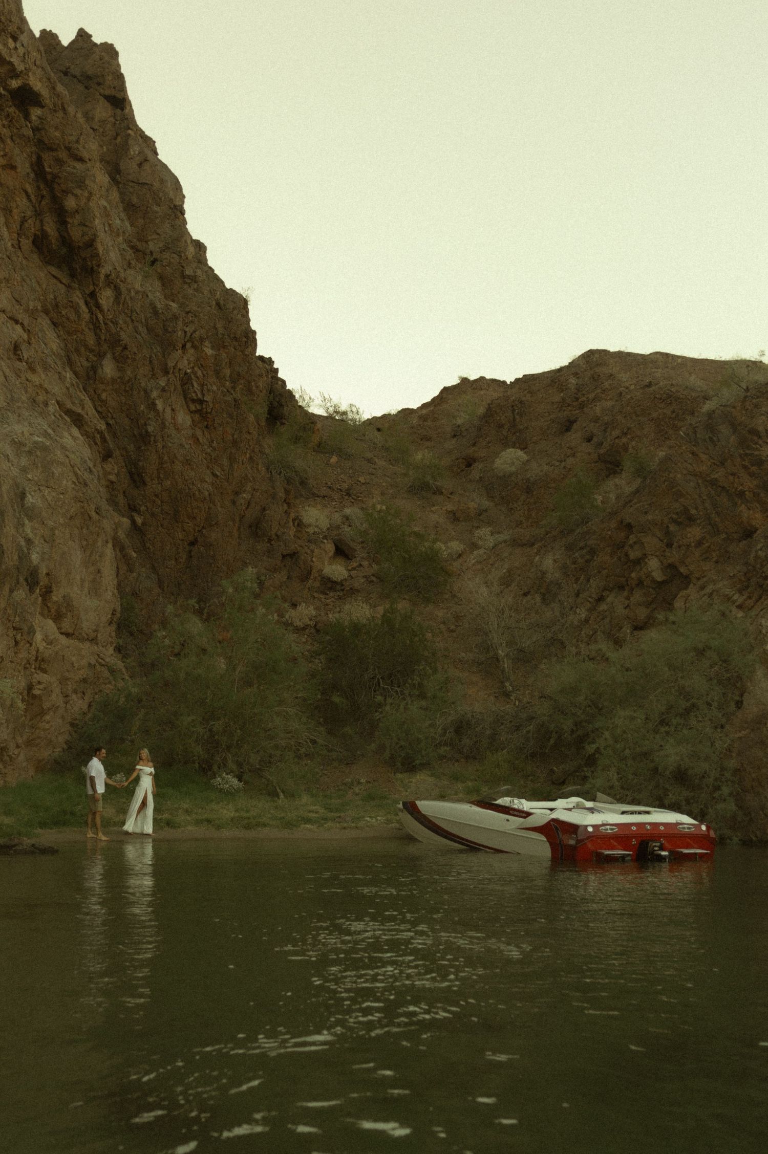 Couple holding hands by a red speedboat on a calm river surrounded by rocky canyon walls.